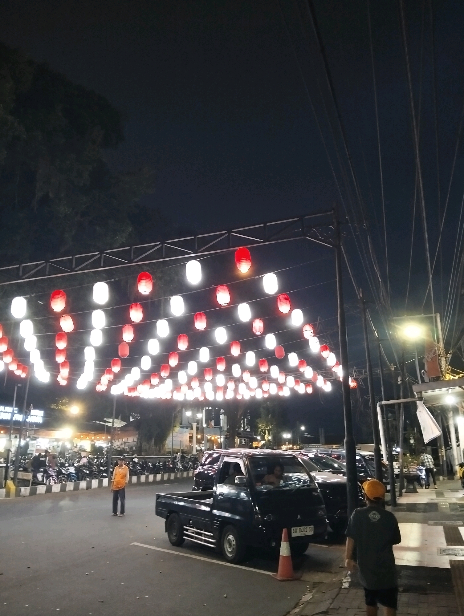 The image depicts a street scene at night in Magelang, Indonesia, likely near a busy area adorned with rows of red and white lanterns strung across wires overhead. In the foreground, a dark-colored pickup truck is parked on the right side of the road, facing left. A person wearing a dark shirt and an orange cap walks away from the camera towards the right side of the frame.
Further down the street, a man in an orange shirt and blue pants stands in the middle of the road, facing the camera. Behind him, a line of motorcycles is parked along the left side of the street, with some people standing near them. Several other vehicles, including cars and possibly more motorcycles, are visible in the background. Buildings with illuminated signs are present on the left side of the street. The overall ambiance suggests a night market or a festive event. The sky is dark, indicating nighttime, and streetlights illuminate the scene.