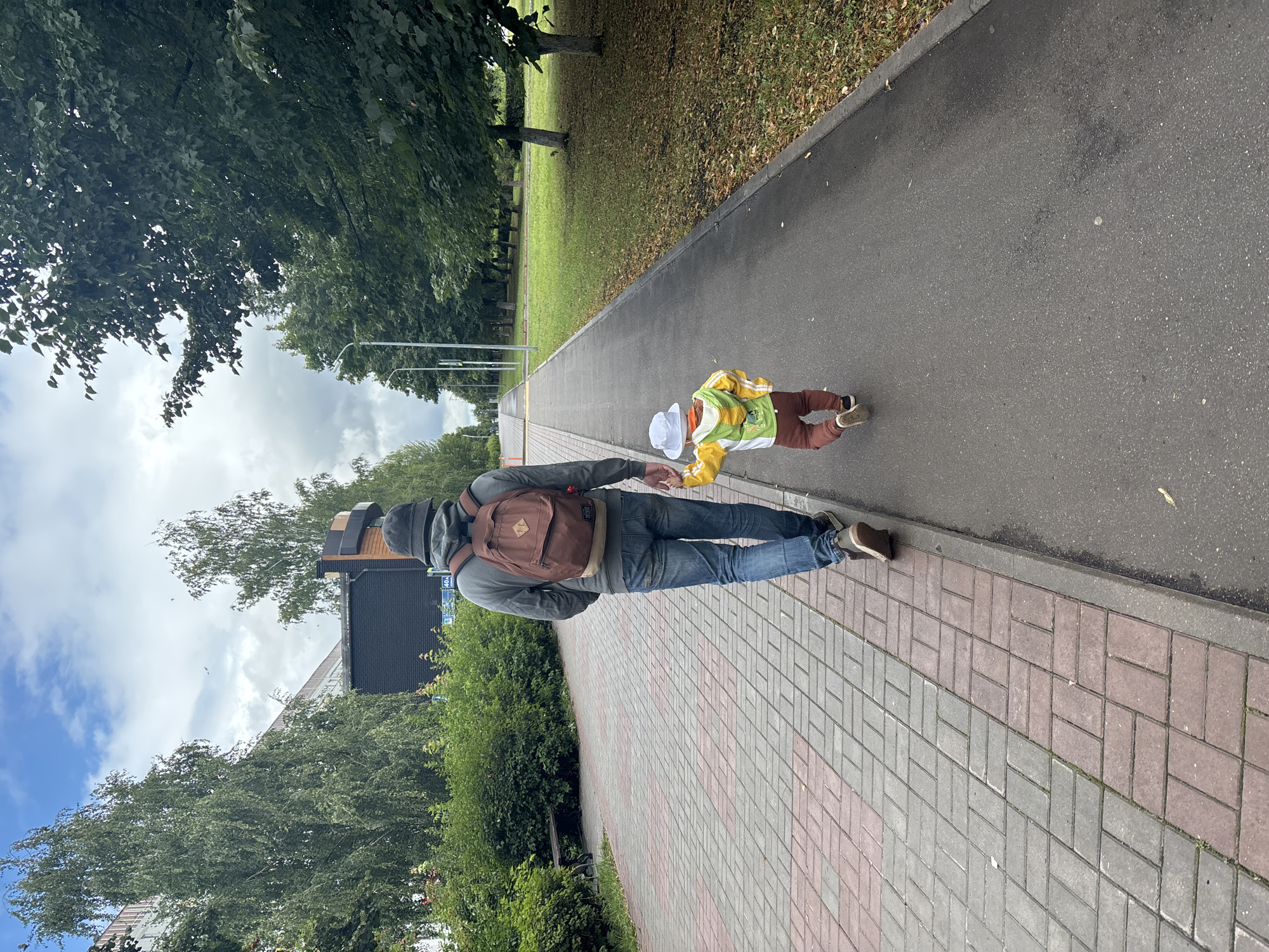 A man and a child are walking away from the camera on a paved path in a park in Naberezhnye Chelny, Russia. The man is wearing a grey hooded sweatshirt, blue jeans, a grey bucket hat, and a brown backpack. He is holding the child's hand. The child is wearing a yellow and green jacket, brown pants, and a white bucket hat. The path is bordered by a grassy area with trees on the right and a brick walkway with bushes on the left. In the background, there are buildings and more trees. The sky is cloudy.