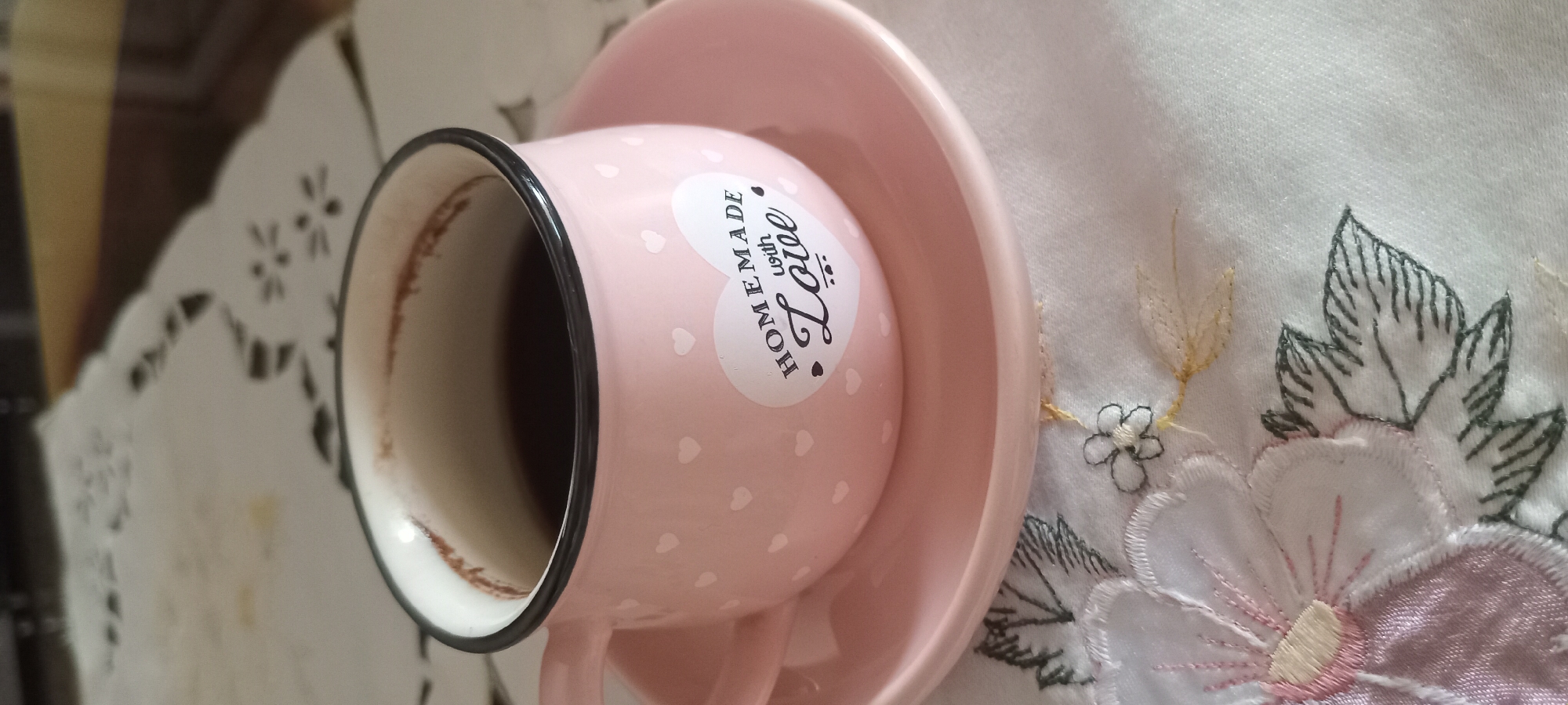 A close-up, eye-level shot shows a pink ceramic coffee cup filled with dark coffee. The cup has a white interior and a thin black rim. A matching pink saucer sits beneath the cup. On the side of the cup, a white sticker with black text reads "HOMEMADE with Love" inside a heart shape. The cup and saucer are placed on a white cloth with intricate embroidery of pink and white flowers and green leaves. The background is blurred, with hints of a light-colored surface and a dark edge.