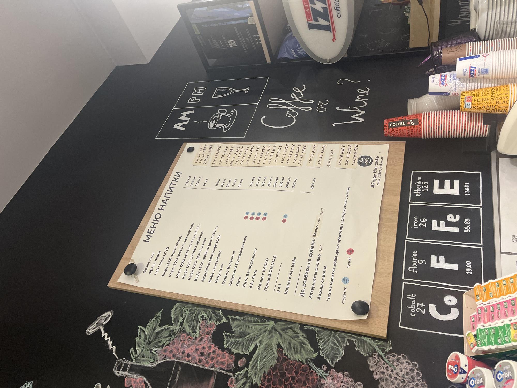 The image presents a close-up view of a menu board at a coffee shop, likely in Plovdiv, Bulgaria, based on the Bulgarian currency (ЛВ) displayed on the menu. The menu lists various hot beverages such as different types of coffee (Frappe, Izzo variations, Americano, Cappuccino), tea, cocoa, and Nescafe with milk, and options for adding milk, alternative milk, or Irish cream. The board includes an "AM PM" graphic alongside the question "Coffee or Wine?". The menu is displayed on a wooden board, with the prices for each item listed in Bulgarian Lev and Euro. A small quantity of Orbit chewing gum can be seen on the right, along with paper cups. The background also has a chalk drawing of a wine bottle with grapes and leaves. The board is partially overlayed with what appears to be a chemical element symbol drawing.