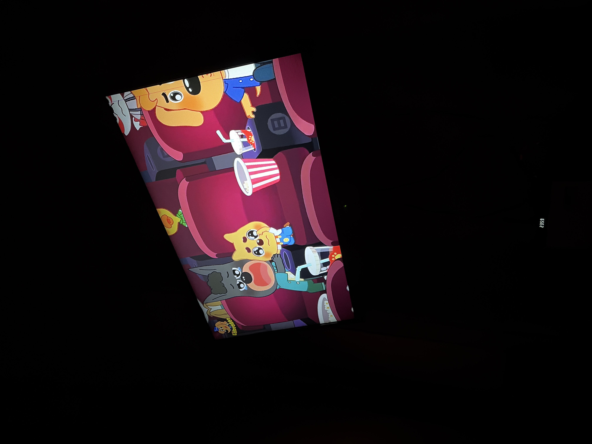The image shows a screen displaying an animated scene of characters in a movie theater. Two cartoon dogs are visible, one appearing distressed and crying, while the other comforts them. They are seated in red theater seats, with popcorn and drinks in front of them. Another cartoon character, a yellow chick, is also present. The screen also displays the text "2359" in the lower right corner, likely indicating a time. The context of Tambov, Russia, is not depicted in the image.