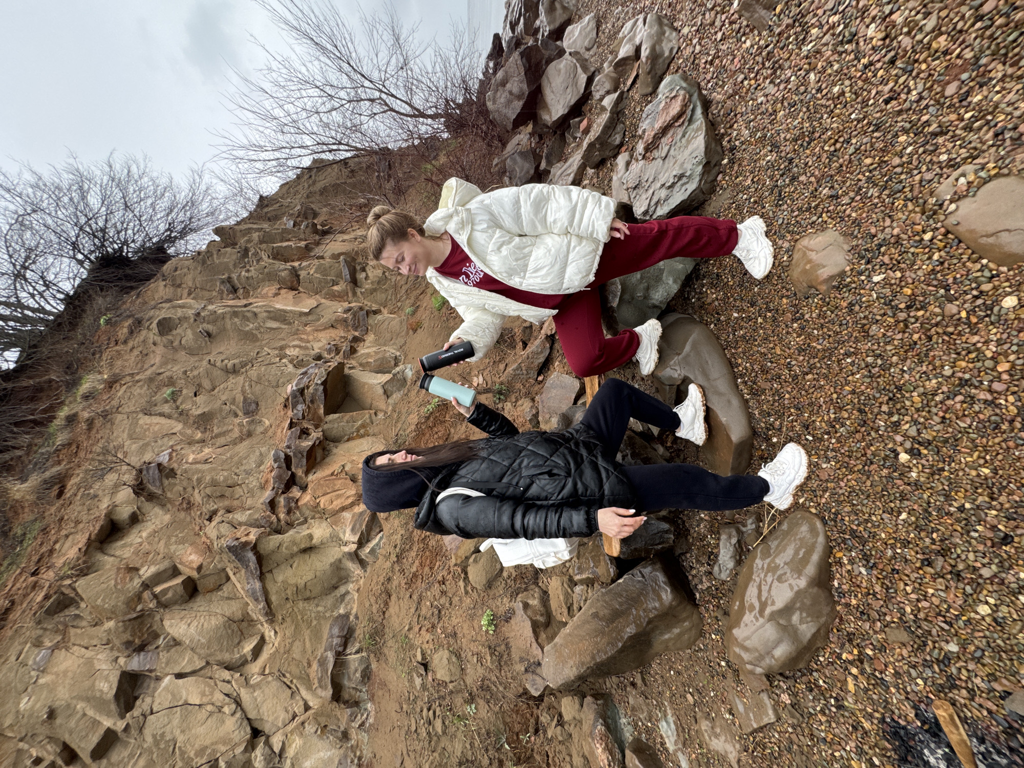 Two women are outdoors in a rocky, hilly environment, possibly a beach or shoreline near Kostovaty, Russia. The woman on the upper right wears a white puffer jacket and maroon athletic pants. She holds a black water bottle in her right hand and a blue water bottle in her left. The woman on the lower left wears a black quilted jacket, black pants, and a black hooded hat. She holds a blue water bottle in her left hand. Both women wear white sneakers. The ground is covered with pebbles, rocks, and damp earth. A cliff face is visible to the left, with bare trees at the top. The sky is overcast.