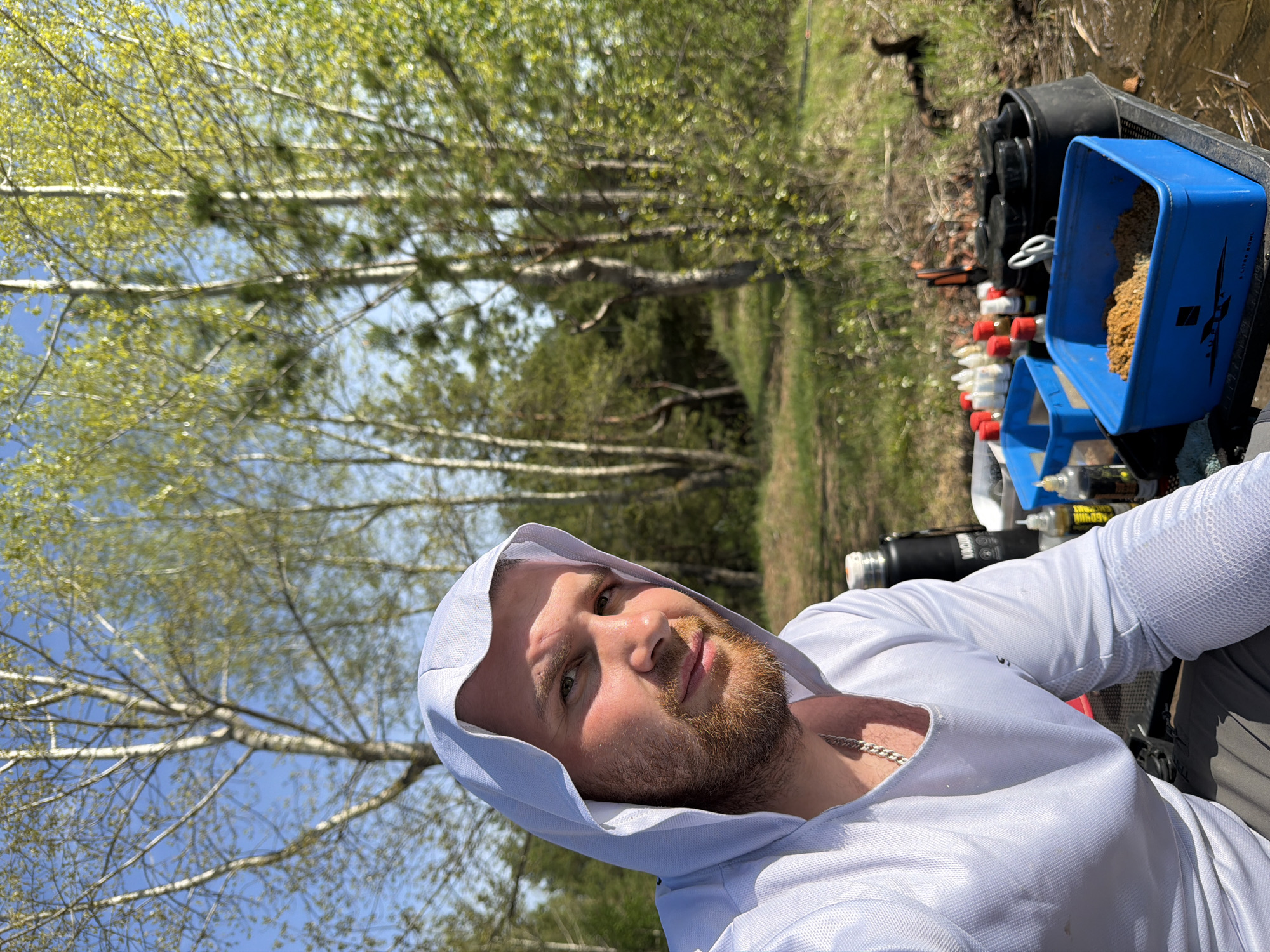 A man wearing a white hooded shirt is outdoors, likely in a natural environment near trees. A blue container filled with granular material and other equipment are positioned to the man's right. The location in Chaykovsky, Russia cannot be confirmed from this image.