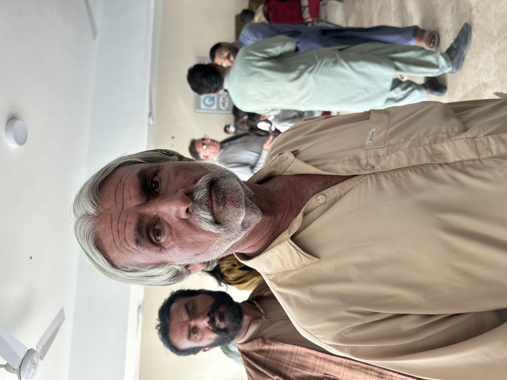 The image depicts a group of men indoors in Quetta City Tehsil, Pakistan. In the foreground, an older man with gray hair and a mustache is looking directly at the camera, wearing a light tan shirt. Behind him, another man with a dark beard is partially visible. Further in the background, several other men are seen standing or sitting, some wearing traditional Pakistani clothing. The environment appears to be a waiting area or office space with light-colored walls and a ceiling fan.