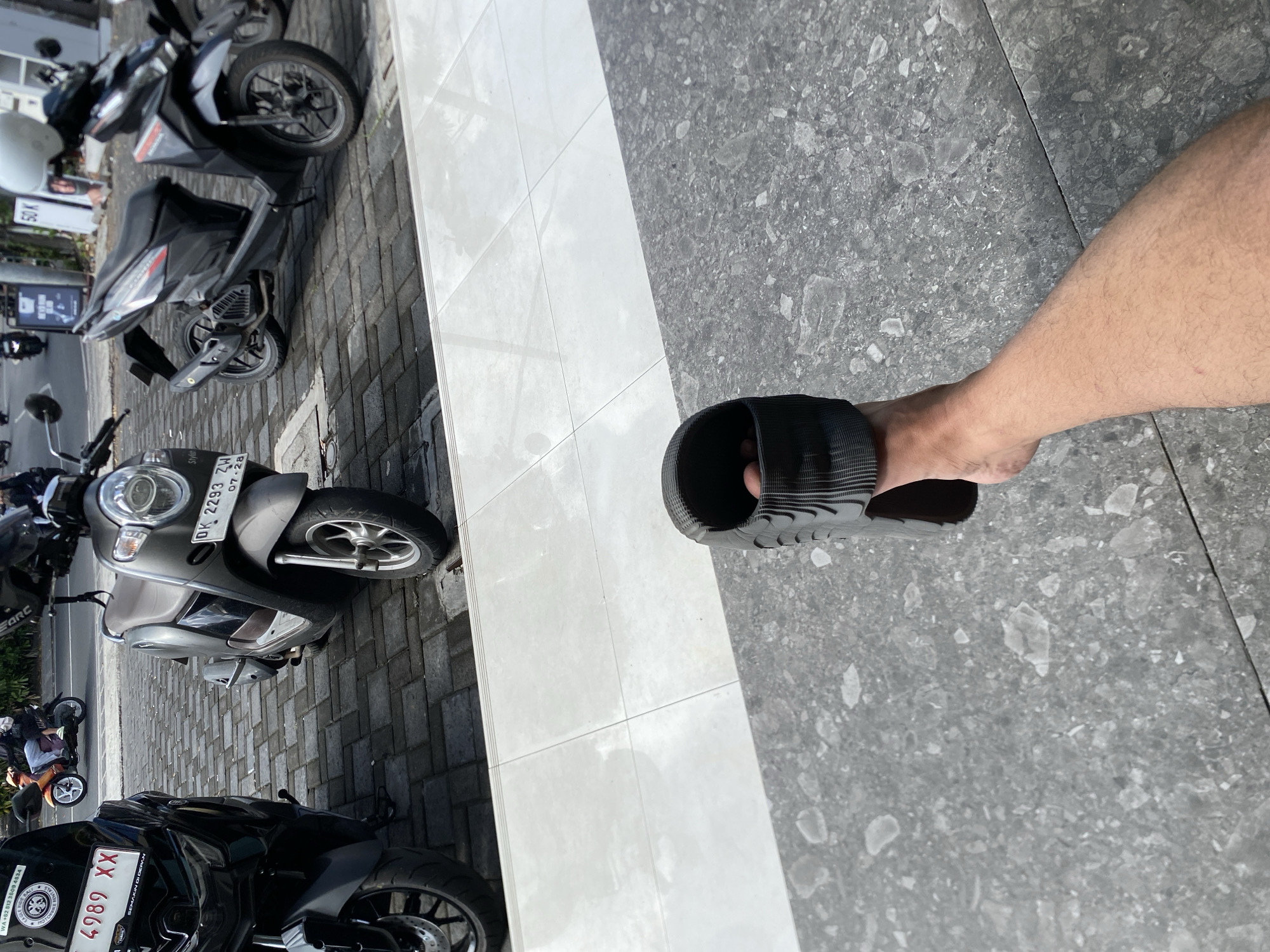 In North Kuta, Indonesia, a person's hand holds a black sandal, with their leg and foot visible in the foreground. The background features several parked scooters and motorcycles on a cobbled street. One scooter has a license plate that reads "DK 2293 ZW" and the date "07-28". Another motorcycle has a license plate that reads "4989 XX". The scene suggests a street-side view with multiple vehicles, possibly in a commercial or busy urban area.