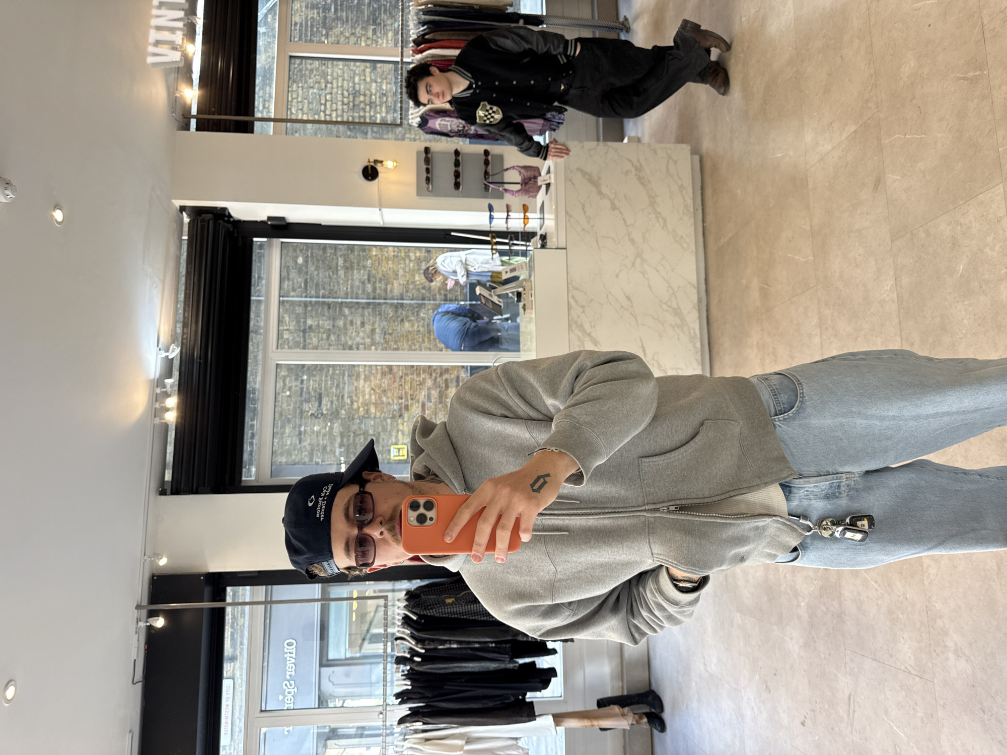 A young man takes a mirror selfie inside a clothing store, which appears to be a vintage or boutique shop. The image, rotated 90 degrees clockwise, captures the man in the foreground holding an orange smartphone, wearing a grey zip-up hoodie, light blue jeans, sunglasses, and a dark baseball cap. Keys on a carabiner clip are visible on his jeans, and he has a tattoo on his right hand.
In the background, reflected in the mirror, another young man stands wearing a black letterman jacket, black pants, and brown shoes. The store interior features clothing racks displaying various garments, a white marble-patterned counter, and display stands showcasing sunglasses and a pink handbag. Through large windows or glass doors, brick buildings are visible outside. The floor is covered with light-colored tiles.
A partially visible, inverted sign reading "VINTAGE" is seen above the mirror. Text on the selfie-taker's baseball cap includes "Dave & Buster's", "Ocean Beach", "Holiday Inn", "Resort", and "Daytona Beach", though partially obscured and mirrored. Other blurry text, "Otlse? revitO", is reflected on a distant window. The scene is brightly lit, suggesting daytime. The overall setting is an urban retail space, consistent with the location context of London, United Kingdom.
