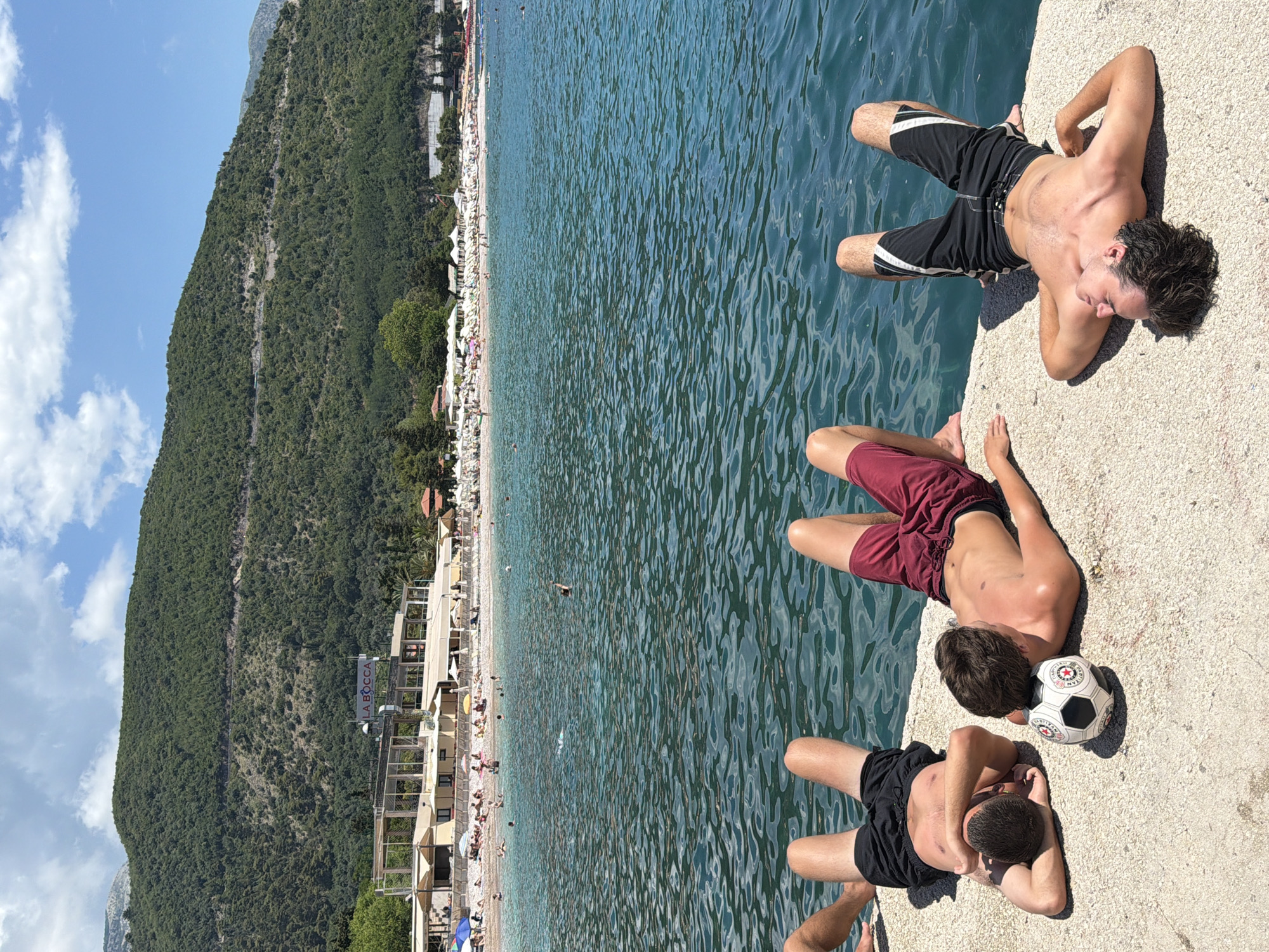 The image shows three young men lying on a concrete ledge overlooking a crowded beach and the Adriatic Sea in Budva, Montenegro. The sun is high in the sky, indicating it is likely midday or early afternoon. The weather appears to be clear and sunny, with a few white clouds scattered across the blue sky.
The man on the far right is lying on his back with his arms behind his head, wearing black and white swim trunks. Next to him, a second man is lying on his side with his head propped up, wearing burgundy swim trunks. In front of him, the third man is also lying on his side, wearing black swim trunks. A soccer ball rests between the second and third men. The men appear relaxed and are enjoying a sunny day at the beach.
The background features a long, pebble beach filled with white beach umbrellas and people sunbathing and swimming in the clear blue water. A lush, green mountainside rises steeply behind the beach. Buildings and some trees are visible along the coastline. A sign with the text "LA BOCCA" is visible on one of the buildings. The overall scene depicts a vibrant summer vacation atmosphere.