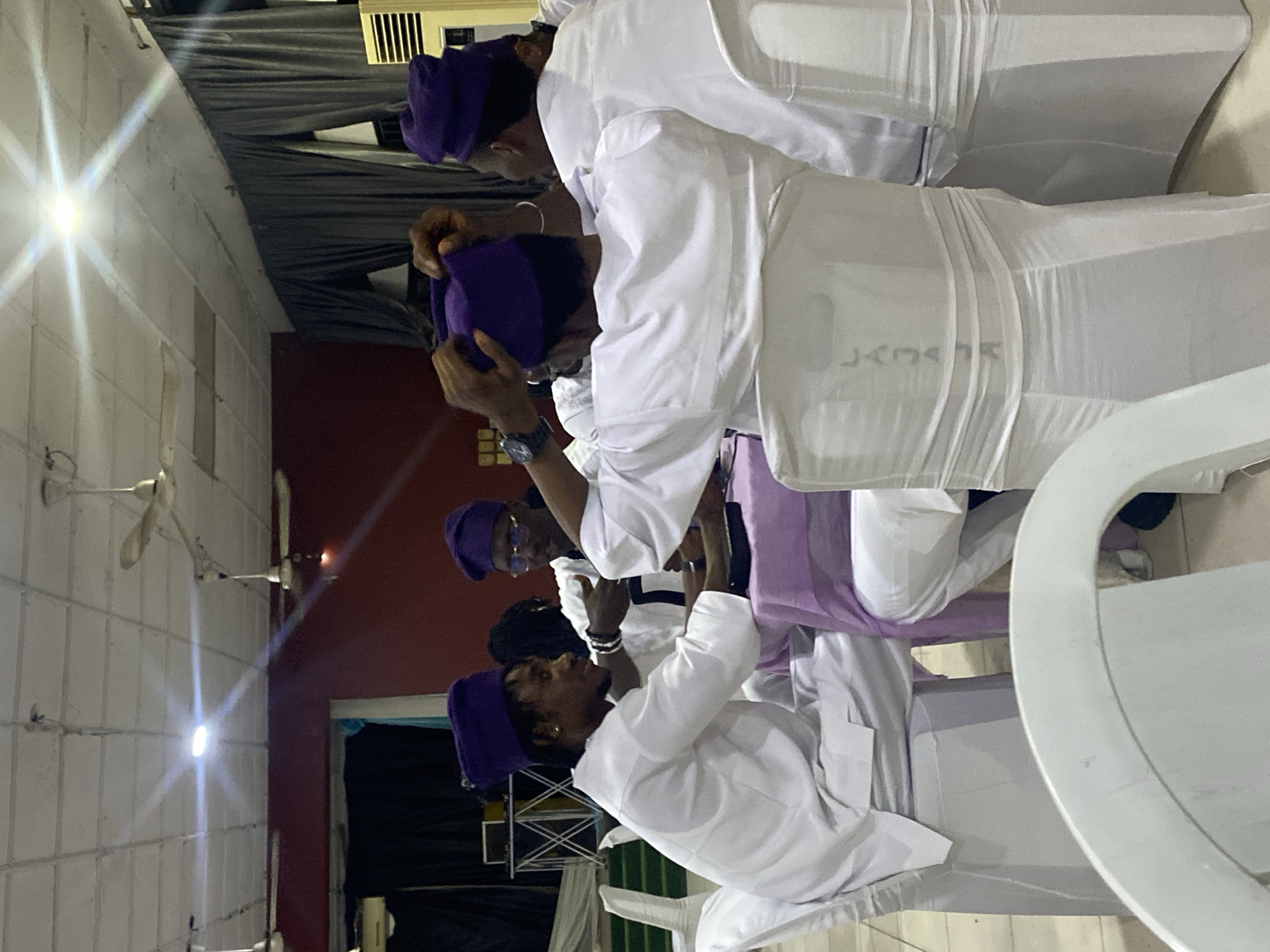 The image shows at least four individuals in white attire, wearing purple caps. They are seated in what appears to be an indoor venue. The environment includes white walls, ceiling fans, and various pieces of furniture. The city and specific location are Unknown. The individuals are adjusting their caps, and the setting seems to be a formal or ceremonial event.