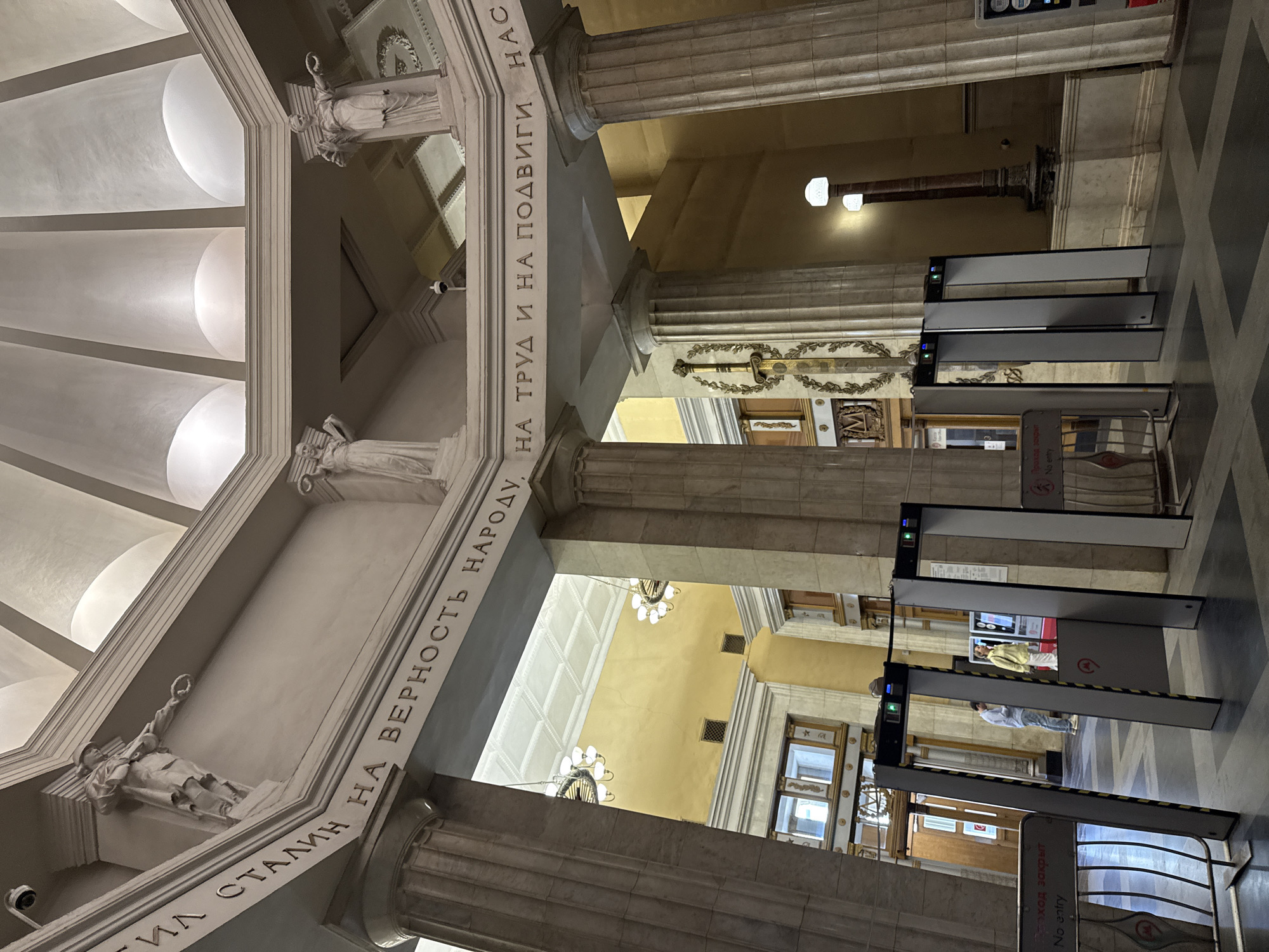 The image depicts the interior of a grand, classical-style hall with a domed ceiling. The walls are adorned with inscriptions and relief sculptures. The inscription reads, "СТАЛИН НА ВЕРНОСТЬ НАРОДУ, НА ТРУД И НА ПОДВИГИ НАС" which translates to "Stalin to the loyalty of the people, to labor and to deeds". Several large, fluted columns support the ceiling. In the foreground, metal detectors are set up, with signs indicating "ВХОД ЗАКРЫТ" and "No entry". Two people, a man and a woman, are visible near the entrance. The woman is wearing a yellow jacket and the man is wearing a blue shirt. The architecture suggests a public building, possibly a museum or a historical monument. Based on the inscription and architectural style, this location is likely the "Palace of Labor" (formerly the Central House of the Red Army) on Leningradsky Prospekt in Moscow, Russia. The scene appears to be a security checkpoint for entry into the building.