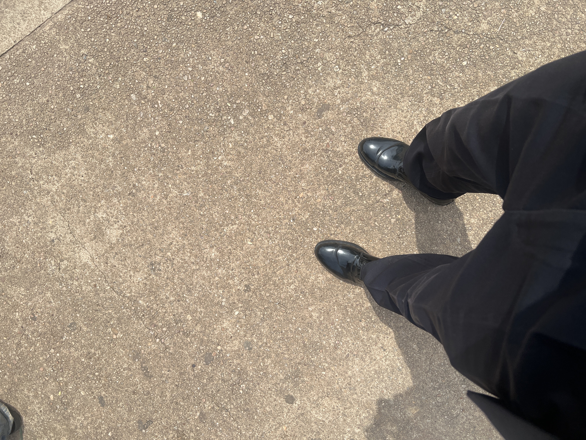 A person is standing on a paved surface, likely a sidewalk or walkway in the city of Ife, Nigeria. The paving is made of aggregate concrete, with visible stones and a rough texture. The person is wearing dark trousers and shiny black dress shoes. The lighting suggests it is daytime, and a shadow is cast to the left of the person, indicating the sun's position. The perspective is from above, looking down at the person's legs and feet.