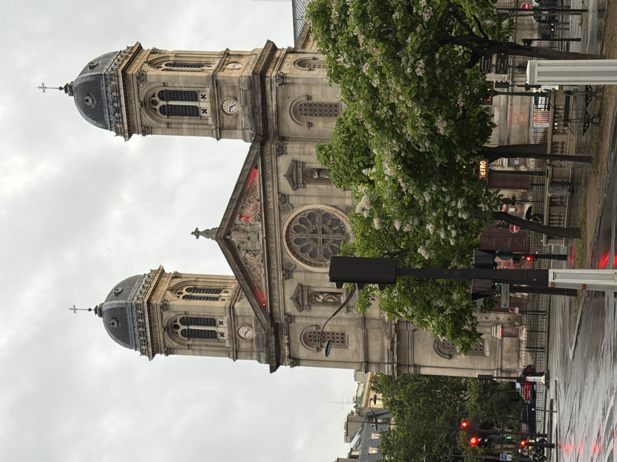 A prominent, ornate church, likely Saint-François-Xavier in Paris, dominates the center of the image under an overcast sky. The church features two symmetrical bell towers, each topped with a dark, domed roof and a cross. The towers are constructed from light-colored stone, adorned with arched windows, decorative carvings, and functional clocks (one visible clock shows approximately 9:30).
The main facade of the church boasts intricate architectural details, including sculptures, decorative pillars, and a large rose window at its center. Above the rose window, a red-lit section highlights a tympanum or pediment, beneath which a statue is visible. Latin inscriptions are carved into the stonework of the church: "HOSANNAH.FILIO.DAVID." on one tower and "GLORIA.IN.EXCELSIS." on the other. Above the rose window, an inscription reads "D.O.M.S.VR.IN H.ET.FRANCISCO.XAVIERIO.A.D.MDCCCXXV," indicating a dedication to Francis Xavier and the year 1825.
Lush green trees with white blossoms partially frame the church on the right side. The ground appears wet, suggesting recent rain. In the foreground and to the right, urban elements are visible, including street lamps, traffic lights (some red, some green), a partially visible glowing "METRO" sign, and railings along a sidewalk. Blurry shapes of vehicles can be discerned on the wet street in the distance. The overall scene depicts a cloudy daytime in an urban environment in Paris, France. No specific human activity is clearly visible.
