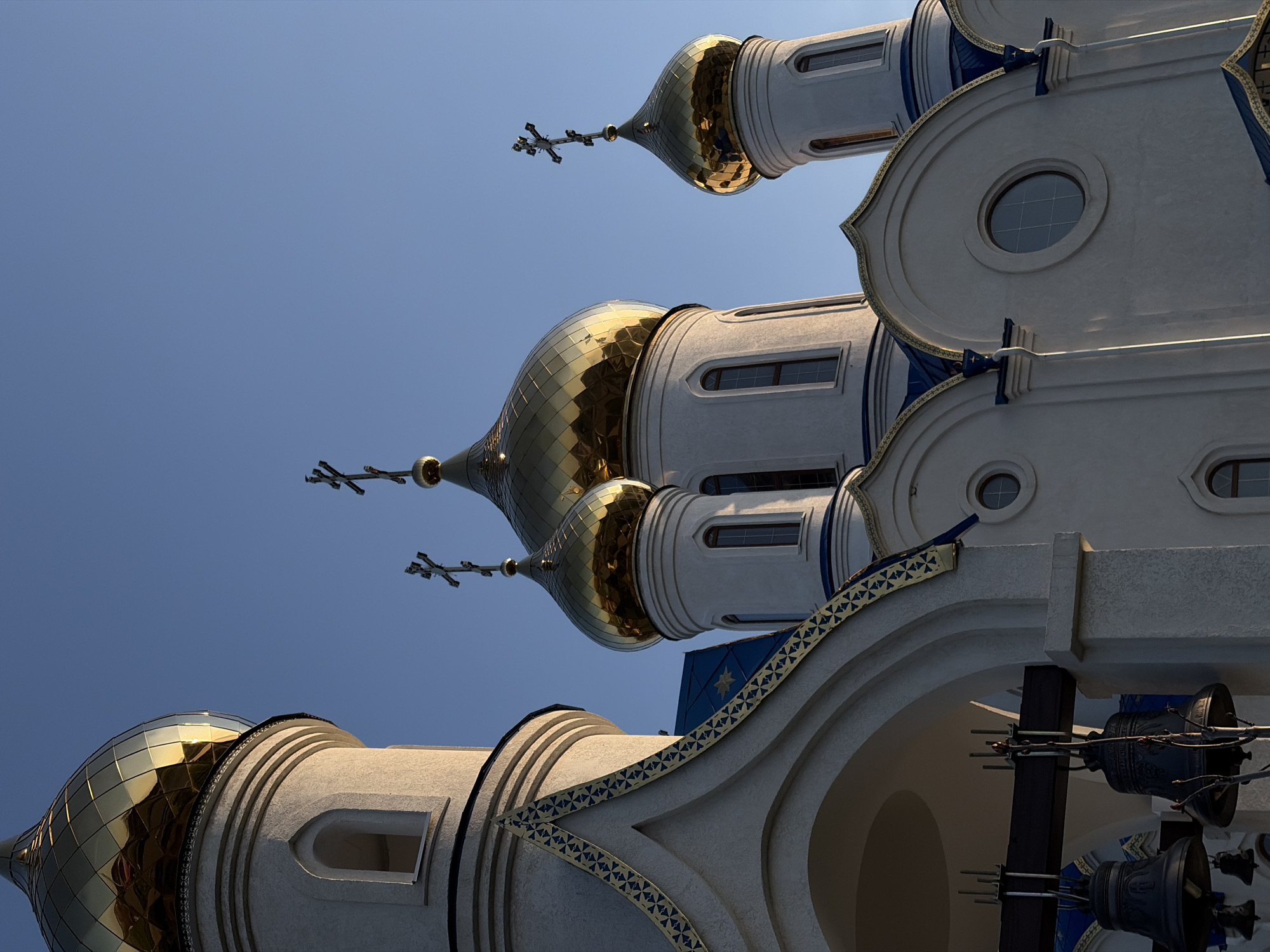 The image showcases the intricate upper architecture of an Eastern Orthodox church or cathedral, viewed from a low-angle perspective against a clear, bright blue sky. Several prominent golden, onion-shaped domes, some featuring a distinct scale-like pattern, reflect the sunlight vividly. Each dome is topped with an elaborate Orthodox cross.
The building's structure is primarily white or light-colored, characterized by graceful curves, arches, and various architectural embellishments. Notable details include circular and arched windows, alongside decorative bands and friezes in striking blue and gold patterns, often incorporating geometric shapes or star motifs. In the lower right of the composition, large, dark bells are visible hanging within an arched opening, suggesting a belfry. The strong, natural lighting and cloudless sky indicate a sunny day. No people or visible text are present. The architectural style is consistent with the provided location context of Novopokrovskaya, Russia.