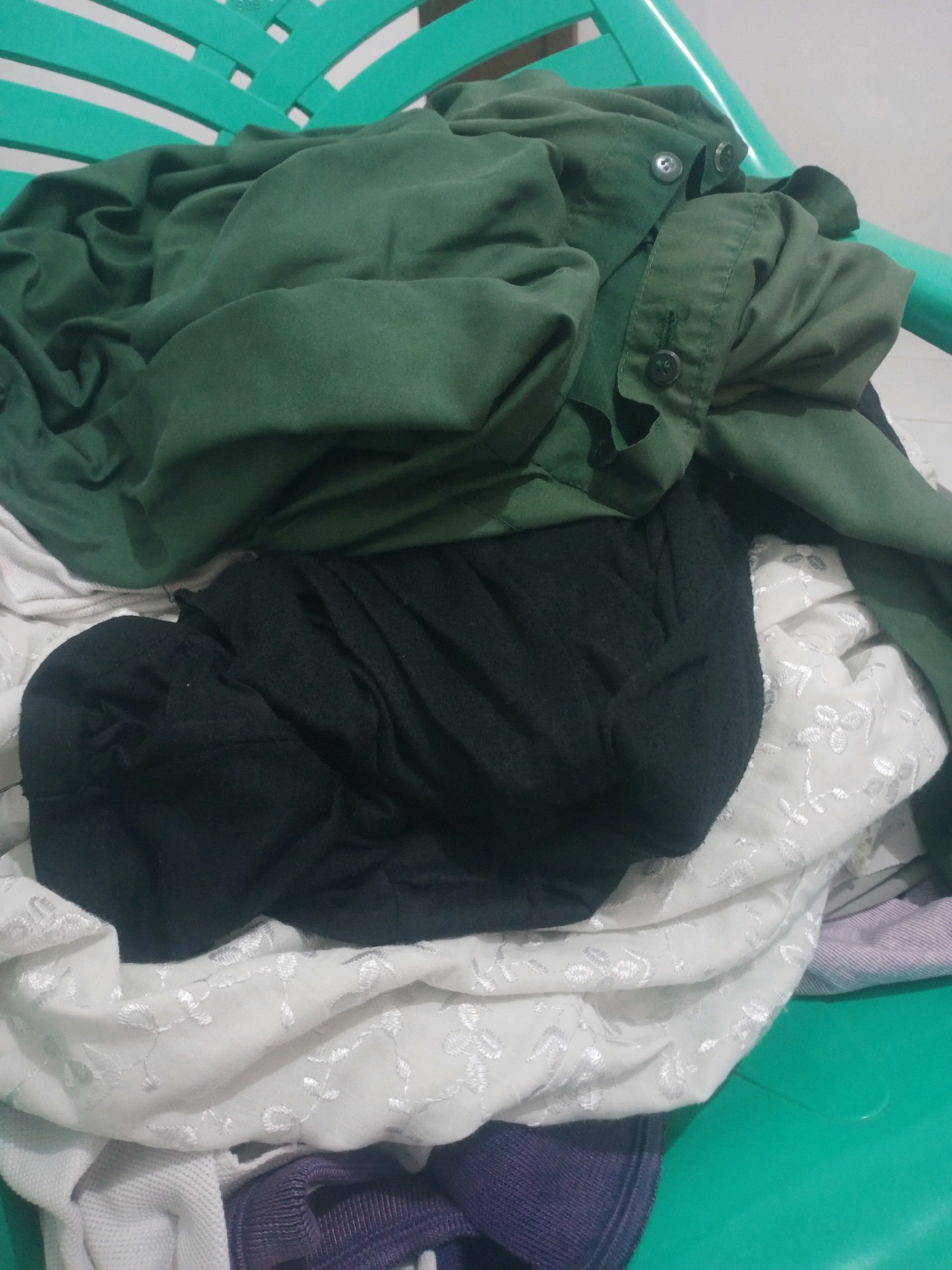 A pile of laundry, including a green button-up shirt, a black t-shirt, and a white embroidered fabric, is resting on a green plastic chair. The scene appears to be indoors, with a white wall visible in the background. The overall impression is one of domestic tidiness or disarray, depending on context. The location in Bandar Lampung, Indonesia, is not visually evident.