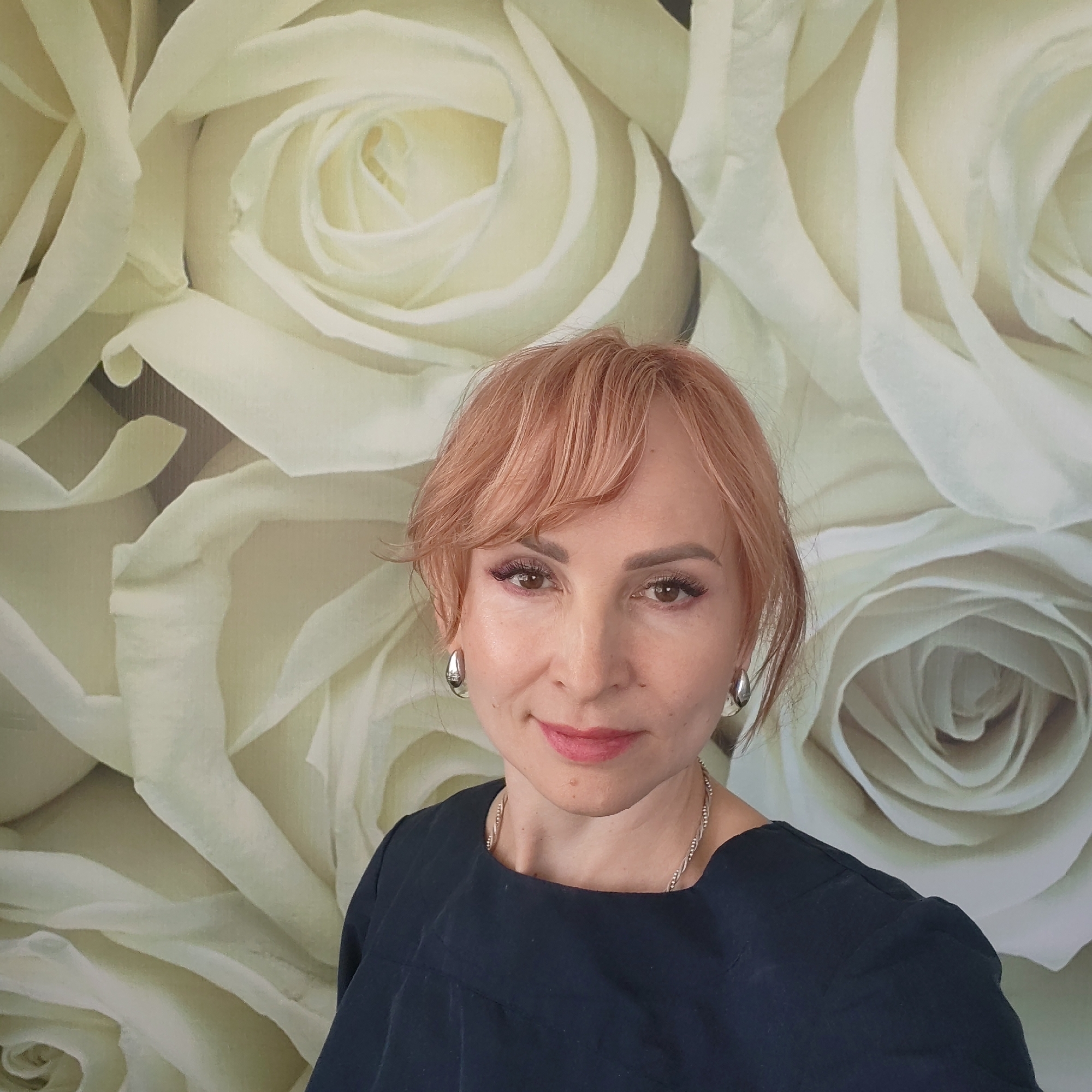 A middle-aged Caucasian woman with short, light reddish-blonde hair, is centered in the frame against a background of large, white rose designs. She has fair skin, brown eyes, and wears subtle makeup. She is wearing teardrop-shaped silver earrings and a silver necklace. She is wearing a dark blue, square-necked top. The location in the city of Chaikovsky, Russia, is not apparent from the image provided.