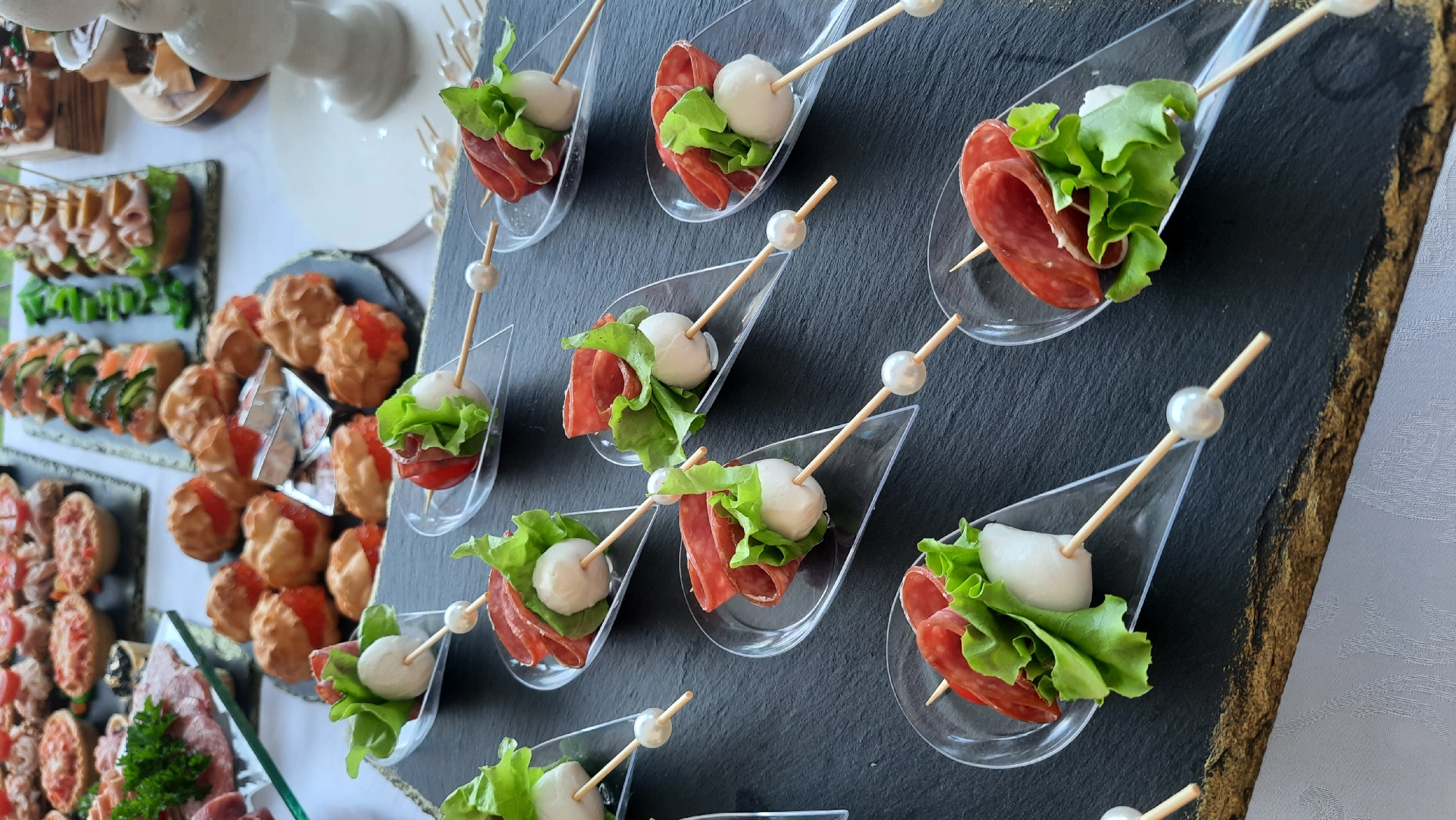 A close-up, high-angle shot showcases an assortment of appetizer skewers arranged on a dark, textured surface, likely a slate platter. The skewers are presented in clear, spoon-shaped serving dishes, each holding a bite-sized creation. These consist of a skewer threaded with a small, white mozzarella ball, a rolled slice of cured salami, and a vibrant green lettuce leaf, often topped with a cherry tomato. The skewers are adorned with small, pearl-like beads. In the background, out of focus, are other appetizers, including small tartlets and what appear to be choux pastry puffs filled with red caviar, suggesting a catering event or a buffet setting. The overall presentation is neat and organized, highlighting the individual appetizer portions.