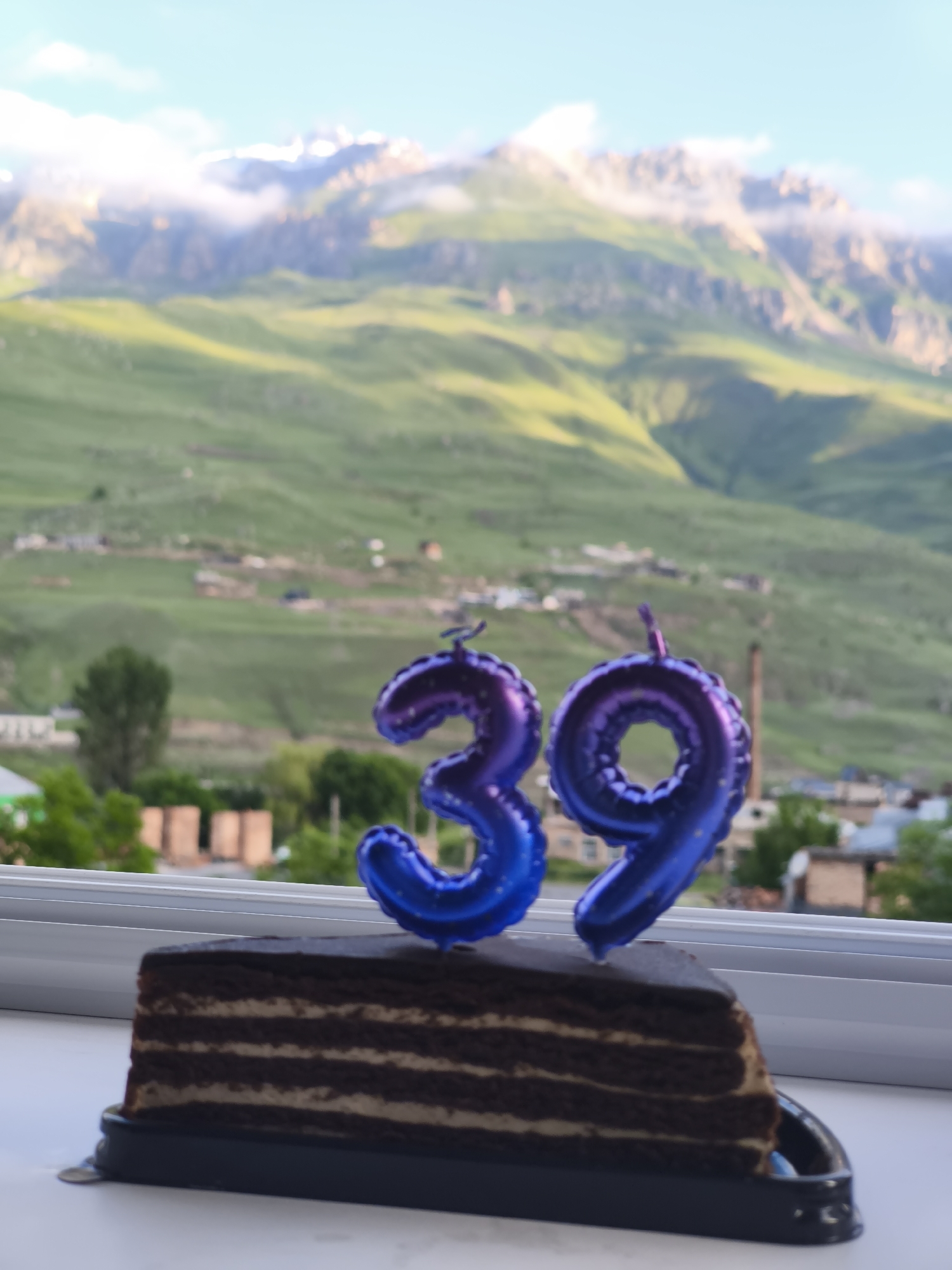 In Verkhny Fiagdon, Russia, a slice of chocolate cake sits on a windowsill, adorned with two purple foil balloons shaped like the numbers "3" and "9". The cake is dark brown with visible layers of cream filling and a glossy chocolate frosting on top. The balloons are inflated and have small strings attached, suggesting they were recently placed.
Beyond the windowsill, a panoramic view of a mountainous landscape unfolds. Rolling green hills, partially obscured by soft clouds, stretch towards jagged, snow-capped peaks in the distance. The sky is a pale blue with scattered white clouds. In the mid-ground, a village with scattered houses and trees is nestled on the slopes of the hills. The overall impression is one of a celebration set against a dramatic natural backdrop.