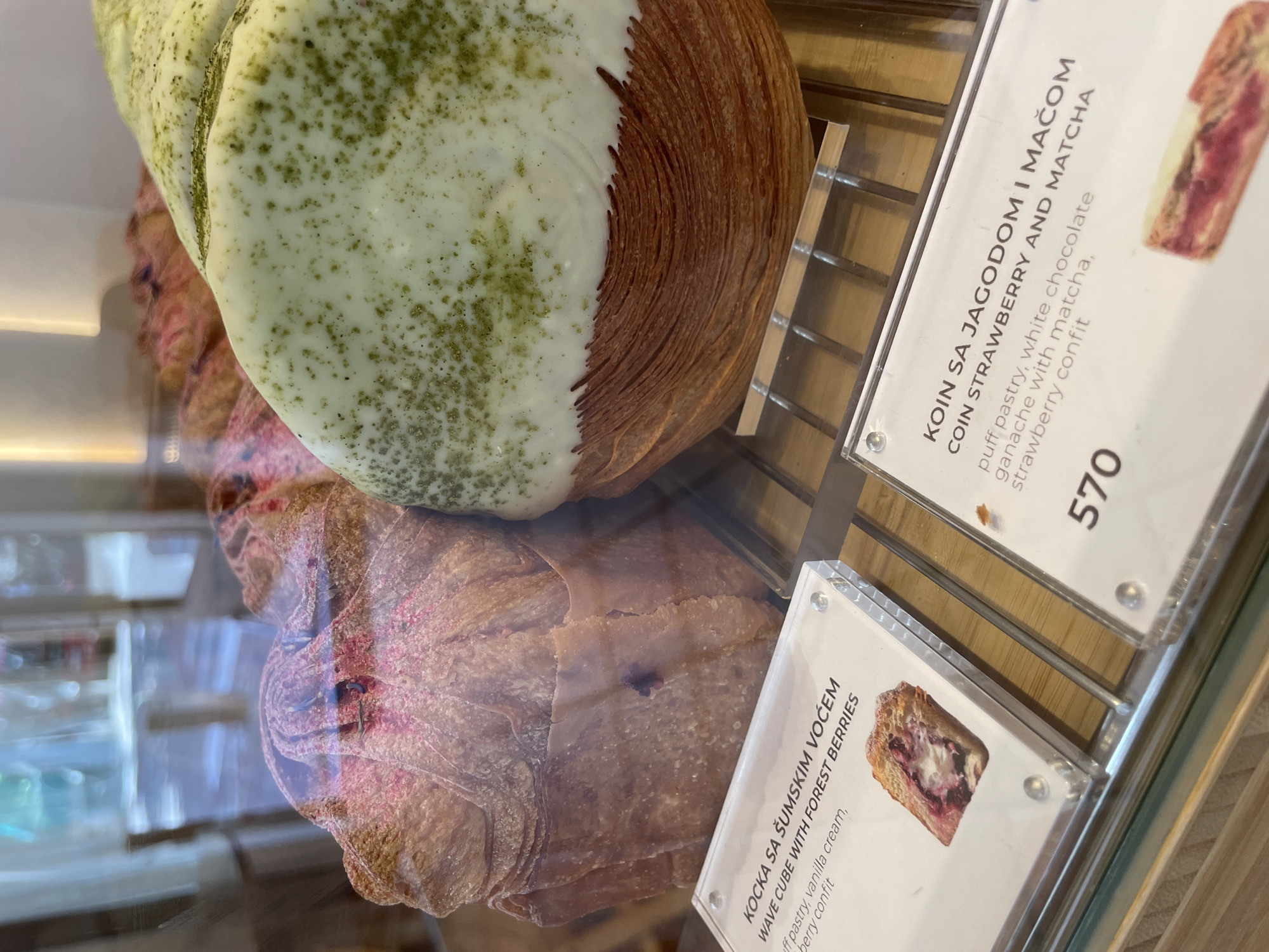 The image shows a close-up of pastries displayed behind glass in a bakery. In the foreground, there are two clear acrylic signs with food descriptions and small pictures of the pastries. The sign on the left reads "KOCKA SA ŠUMSKIM VOĆEM WAVE CUBE WITH FOREST BERRIES puff pastry, vanilla cream, berry confit". The sign on the right is partially visible and reads "KOIN SA JAGODOM I MAČOM COIN STRAWBERRY AND MATCHA puff pastry, white chocolate ganache with matcha, strawberry confit" and below that the number "570".
The pastries themselves are large, layered, and appear to be some type of croissant or danish. The pastry on the left has a pinkish hue and appears to have visible berry pieces. The pastry on the right is topped with a light green glaze, speckled with darker green specks, likely matcha, and a white glaze underneath. Both pastries are on a metal rack behind the glass display.
The background is blurred, showing reflections of an indoor environment, possibly a cafe or shop, with bright lighting and some indistinct furniture or decorations. The overall impression is a clean, modern bakery display case. The presence of Serbian text on the signs strongly suggests the location is Belgrade, Serbia.