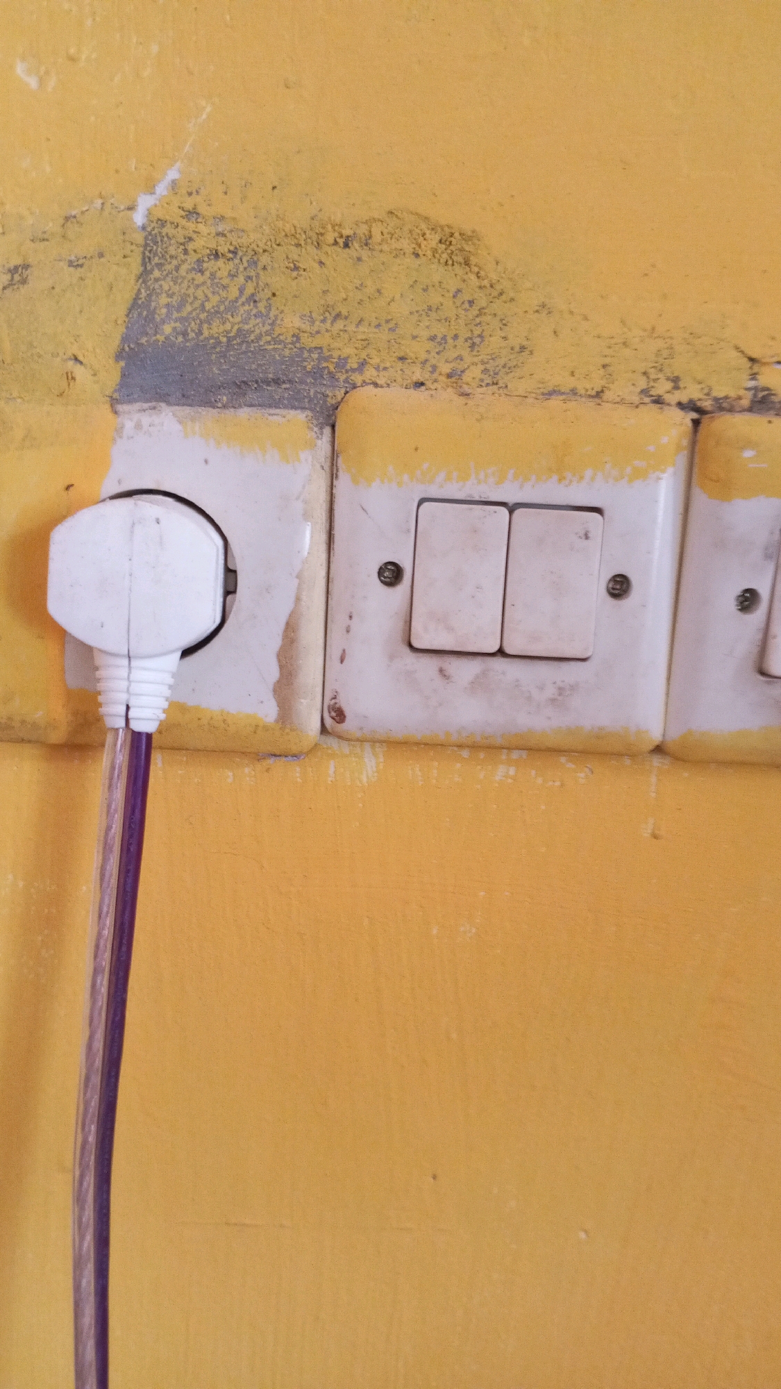 The image shows a close-up of a yellow wall with a cluster of electrical outlets and light switches. One white electrical plug is inserted into an outlet on the left. A purple-sheathed electrical cable extends downwards from the plug. To the right of the outlet, there are two light switches mounted side-by-side on a white plate. Further to the right, another outlet plate is visible, partially obscured. The wall surface exhibits signs of wear, with some peeling paint and discoloration, particularly around the electrical fittings. The overall impression is one of a functional but aged electrical installation in an indoor setting. The location is stated to be in the city of Sritabaang, Indonesia. There are no people visible in the image.