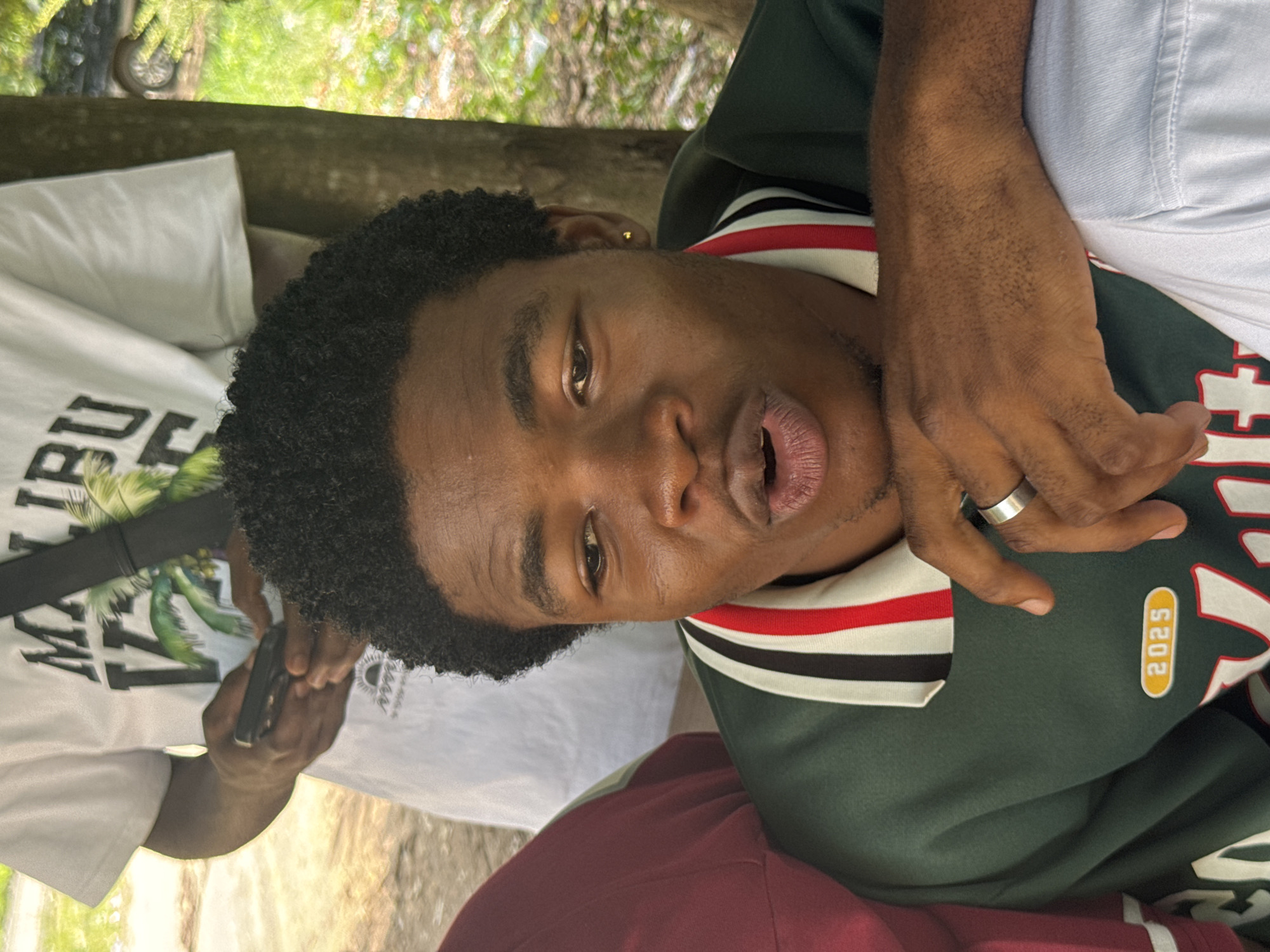 The image shows a close-up of a dark-skinned man with short curly hair. He is wearing a green sweater with white, black, and red striped trim at the collar. The sweater also has a "2025" tag on it. He has a silver ring on his left hand. Another person, wearing a white T-shirt with the word "MALIBU" visible and holding a black cell phone, is partially visible on the left. The background shows greenery, including trees. The location is identified as Ihiagwa, Nigeria.
