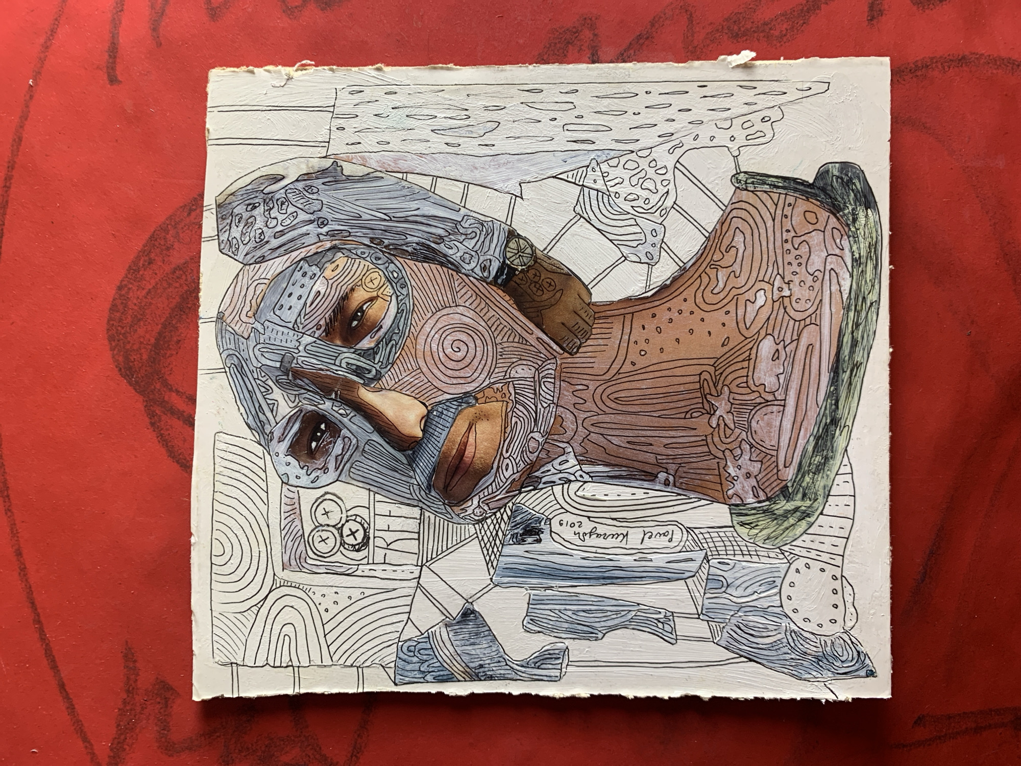 The image is a close-up, overhead shot of a mixed-media artwork featuring a portrait of a person wearing an intricate, patterned mask that covers most of their face. The artwork is on a textured, off-white surface, possibly paper or canvas, with torn edges. The portrait is rendered with linework and colored areas, featuring earthy tones and blues. The background of the artwork is filled with abstract patterns and geometric shapes. The artwork is placed on a vibrant red surface, possibly a table or floor, which has some dark, scribbled markings on it. The artwork appears to be signed "Pavel Krasnograd" with the year "2019". There is no discernible information within the image to indicate the location in Sergeevo, Russia.