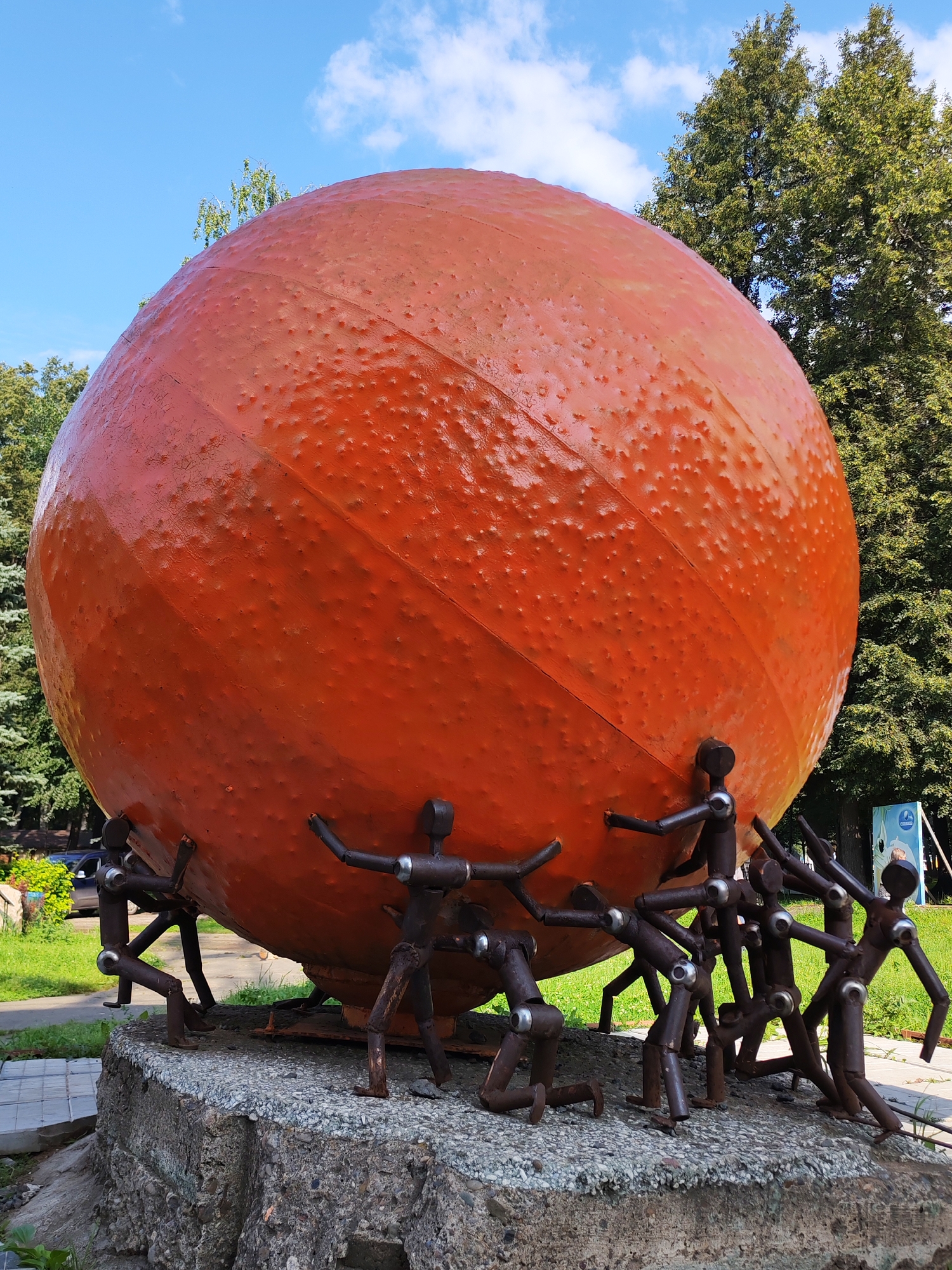 An outdoor sculpture depicts a large, orange, spherical object, textured to resemble a ball, being supported by numerous small, dark, humanoid metal figures. These figures are posed as if lifting or pushing the sphere from below, with many bent at the knees or leaning forward. The sculpture rests on a rough, concrete pedestal. The background features green trees and a blue sky with scattered clouds. A sign with blue and white graphics is visible in the far right background. This sculpture is located in Izhevsk, Russia, specifically on Pushkin Street, near the National Museum of Udmurtia.