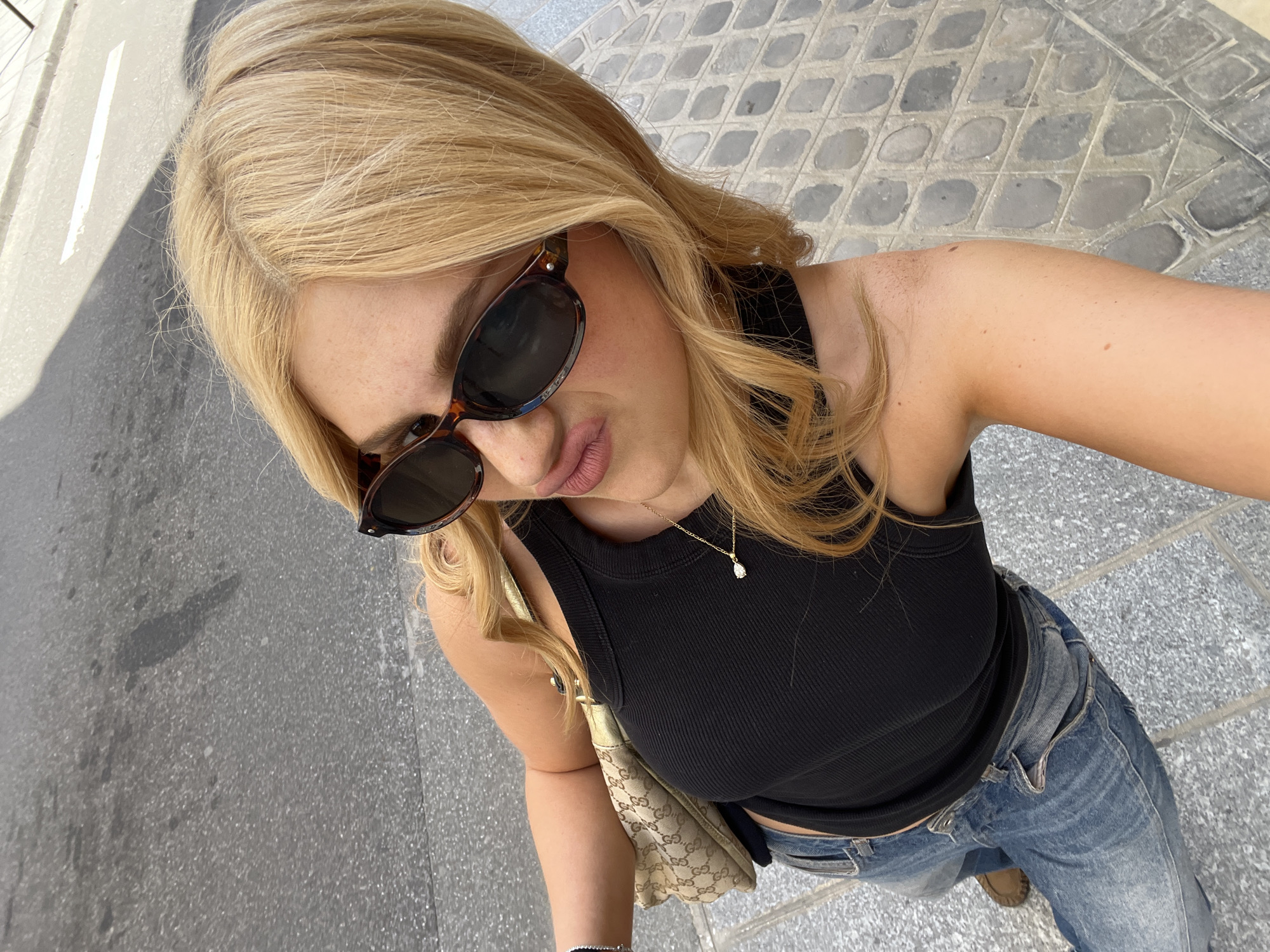 A selfie taken at a downward angle shows a woman with blonde hair and sunglasses standing on a stone-paved sidewalk in Paris, France. She is wearing a black tank top, jeans, and a gold necklace with a pendant. She has a Gucci bag slung over her shoulder. The sidewalk is composed of light grey stone tiles with darker grey grout. A narrow strip of asphalt is visible on the left edge of the frame.