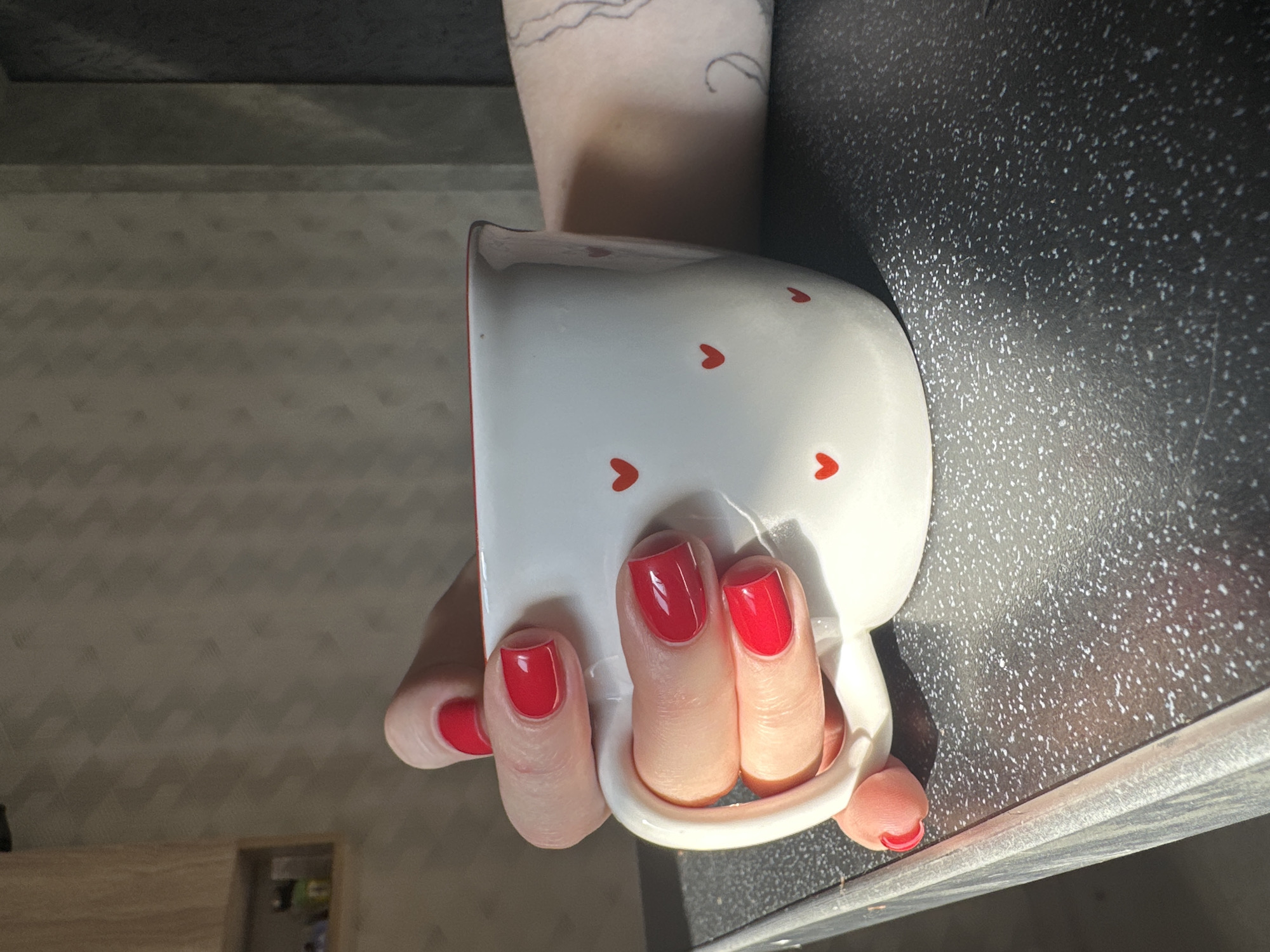 A hand with red nail polish holds a white mug decorated with red hearts. The background includes a dark countertop and a glimpse of a wall. There is no indication of a location in the city of Chaikovsky, Russia.