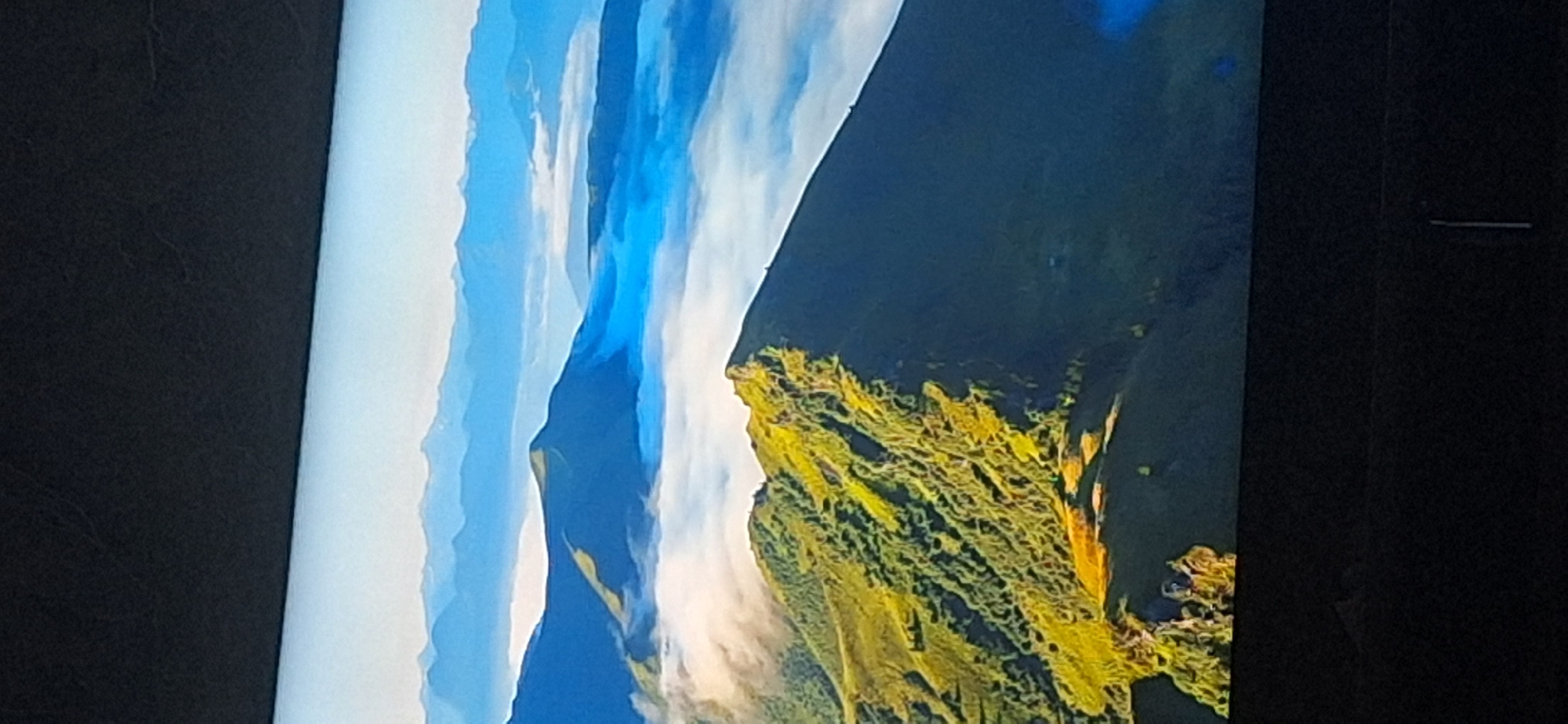 The image depicts a vast mountain landscape with layers of peaks and valleys. In the foreground, a steep, grassy mountain ridge is illuminated by sunlight, casting shadows. The ridge is covered in green vegetation, with visible trees or bushes. Further in the distance, a sea of white clouds fills the valleys between mountain ranges, creating a dramatic effect. Beyond the clouds, more mountain peaks are visible, fading into a hazy blue sky. The scene conveys a sense of immense natural beauty and tranquility. The image is a photograph, possibly taken from a high vantage point, capturing the grandeur of the mountains and the ethereal quality of the clouds. There is no visible indication of the city of Bila Tserkva, Ukraine in this image.