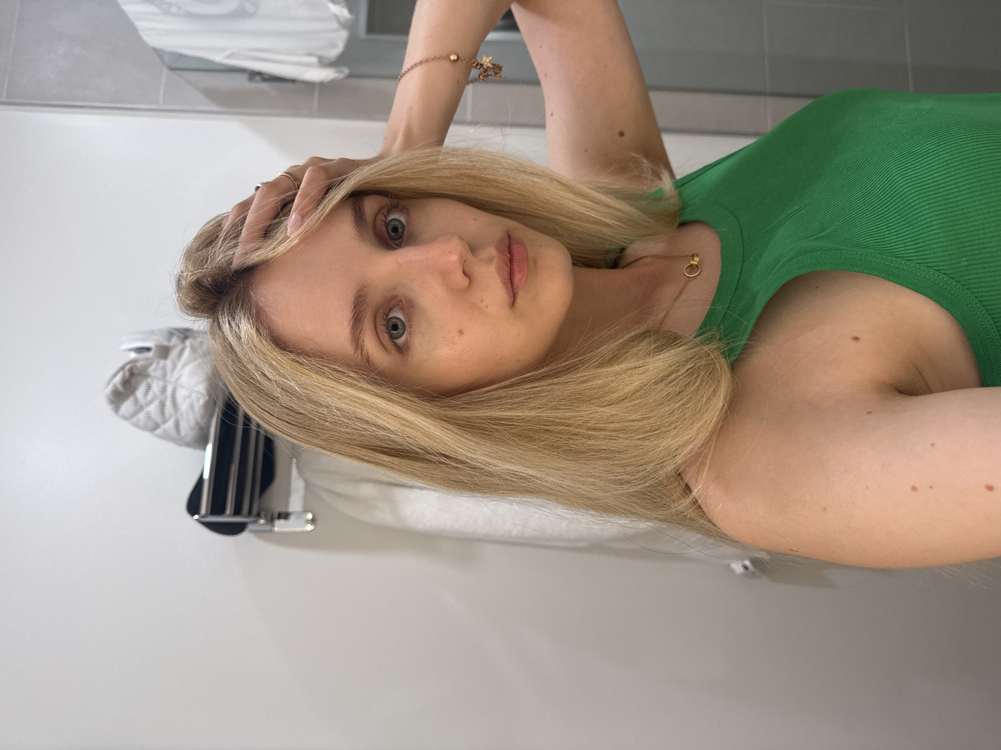 A young woman with blonde hair and blue eyes is taking a selfie in a bathroom. She is wearing a green ribbed tank top and a gold necklace with a small circular pendant. Her right arm is raised and her hand is resting on her head, with a ring on her index finger. A bracelet with a charm is visible on her left wrist. She has fair skin with freckles and a mole on her right shoulder and another on her left arm. The background appears to be a modern bathroom with light-colored tiles on the wall and a light-colored countertop or vanity. A white quilted item and a metallic object, possibly a towel rack, are visible to the left of the woman. The lighting suggests it is daytime, possibly indoors with artificial light. The woman's expression is neutral, and she is looking directly at the camera. The overall impression is a casual and candid moment captured in a personal setting.