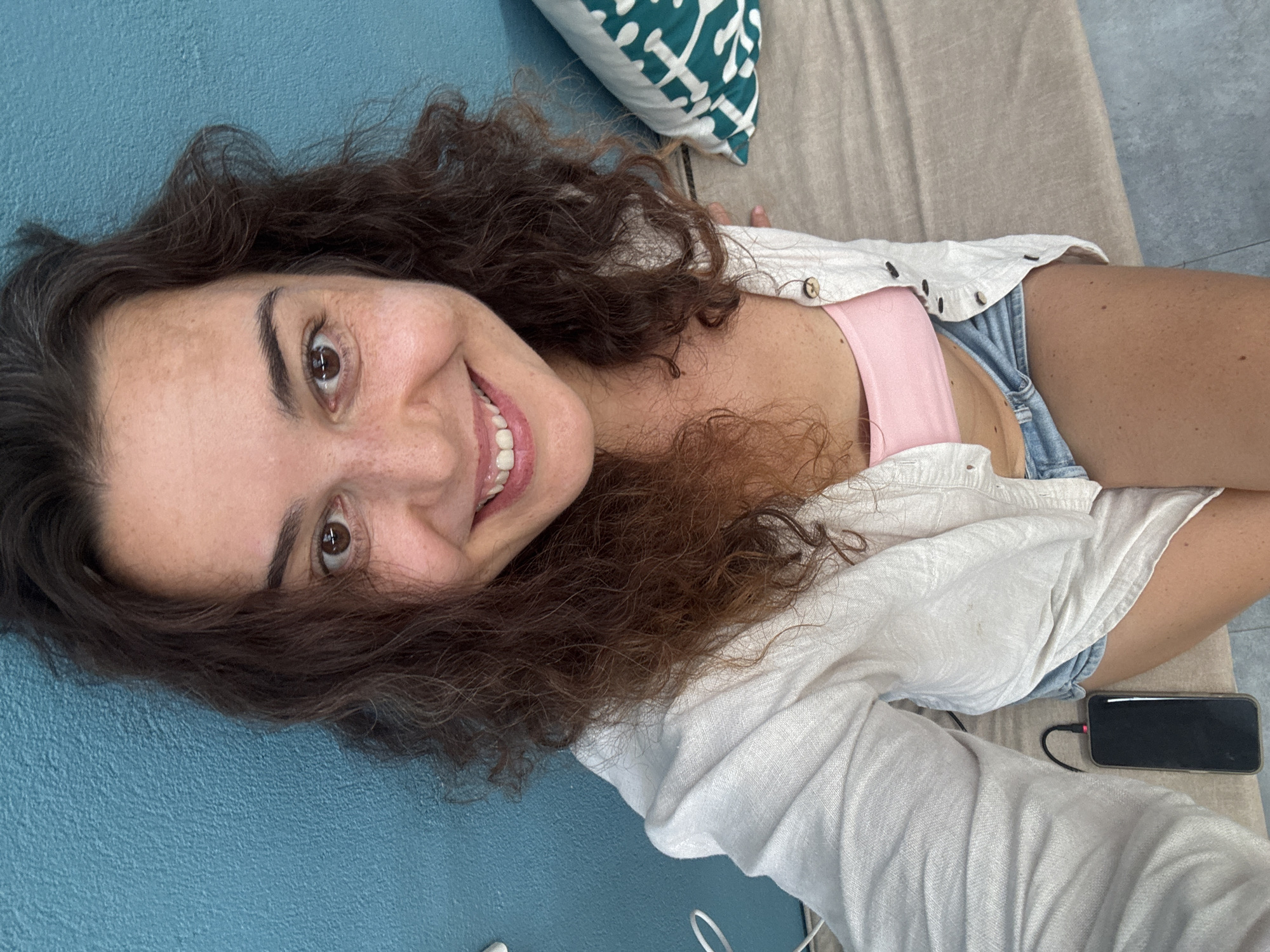 The image shows a woman taking a selfie. She has brown curly hair and freckles on her face. She is wearing a pink tube top, denim shorts, and a white linen shirt. She is sitting on a couch with a blue wall behind her. A phone is visible on the couch next to her. There is also a pillow with a green and white pattern on the couch. Based on the overall aesthetic and the presence of vibrant blue walls, which are common in Hoi An, Vietnam, this scene could be located in a modern cafe or guesthouse in Hoi An.