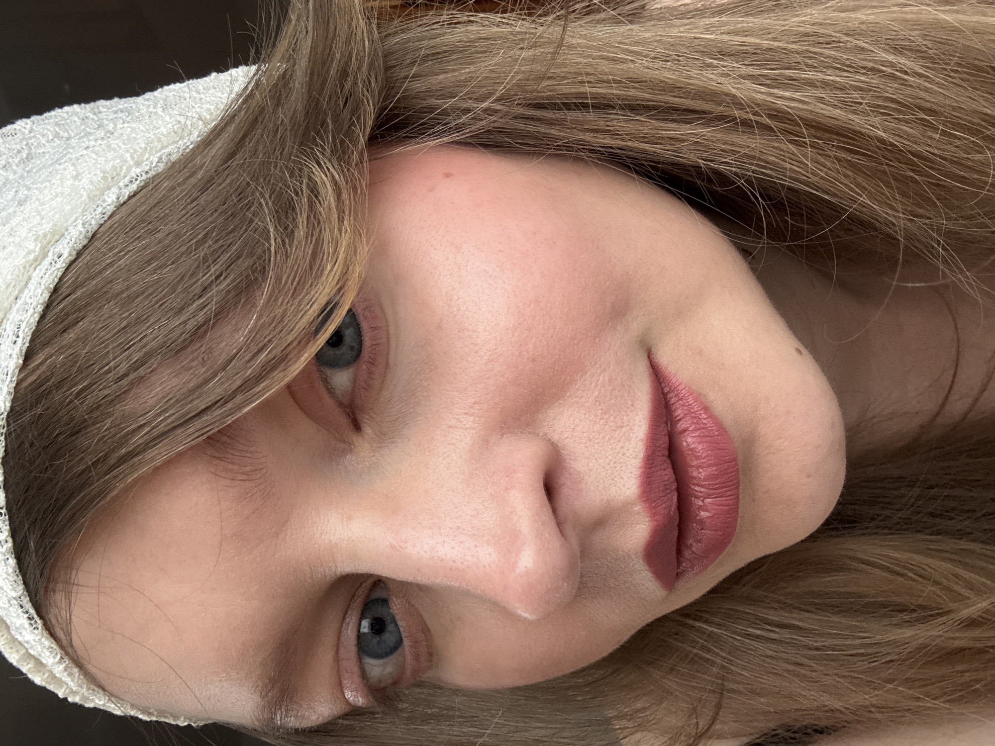 A close-up, rotated view of a woman's face. She has fair skin, blue eyes, and light brown hair with a white lace head covering. She is wearing dark pink lipstick and blush. The background is dark and out of focus. The image provides no information about Izhevsk, Russia.