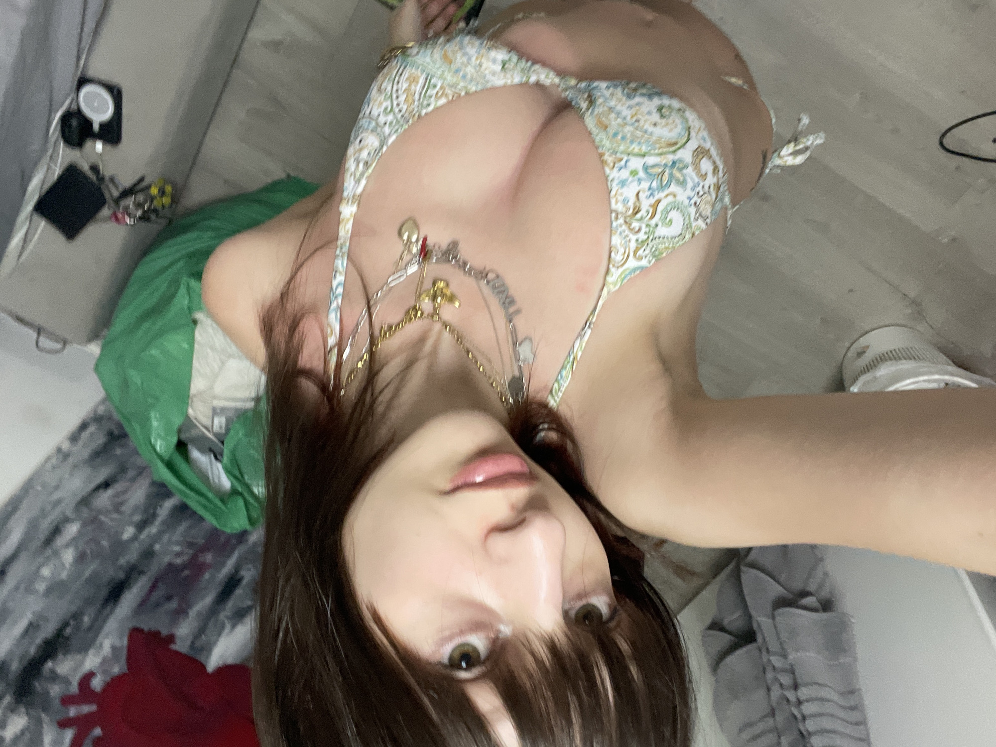 A selfie taken by a female with long brown hair and brown eyes, wearing a paisley bikini top and multiple necklaces, from a high angle looking down at herself. The background includes a bed with grey and red bedding, a green plastic bag, a white shoe on a hardwood floor, and electronic devices including what looks like a charger and smartphone. It is not possible to pinpoint the exact location in the city of Raanana, Israel, with the given information.