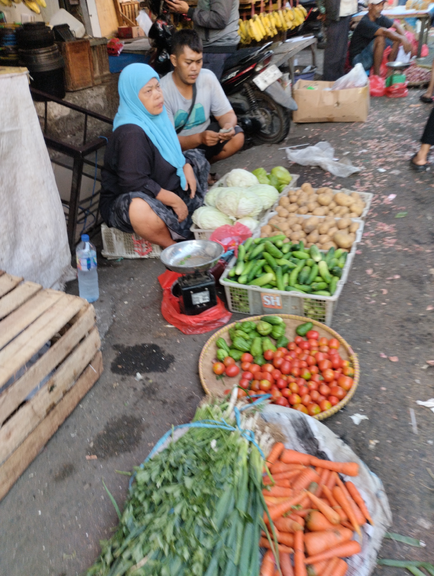 In Bekasi, Indonesia, a street market scene unfolds. A woman wearing a blue hijab and black clothing sits on a crate, her legs crossed, facing right. Beside her, a man in a light grey t-shirt and dark pants also sits. In the foreground, a variety of fresh produce is displayed in baskets and on the ground. Cabbages, potatoes, cucumbers, tomatoes, carrots, and leafy greens are visible. A digital scale sits on a red plastic sheet, with a metal weighing pan. Several plastic bottles, one filled with water, are also present, as is a wooden crate. The ground is paved with asphalt, showing some wet patches and scattered debris. In the background, a motorcycle is parked, and there are glimpses of other market stalls and people.