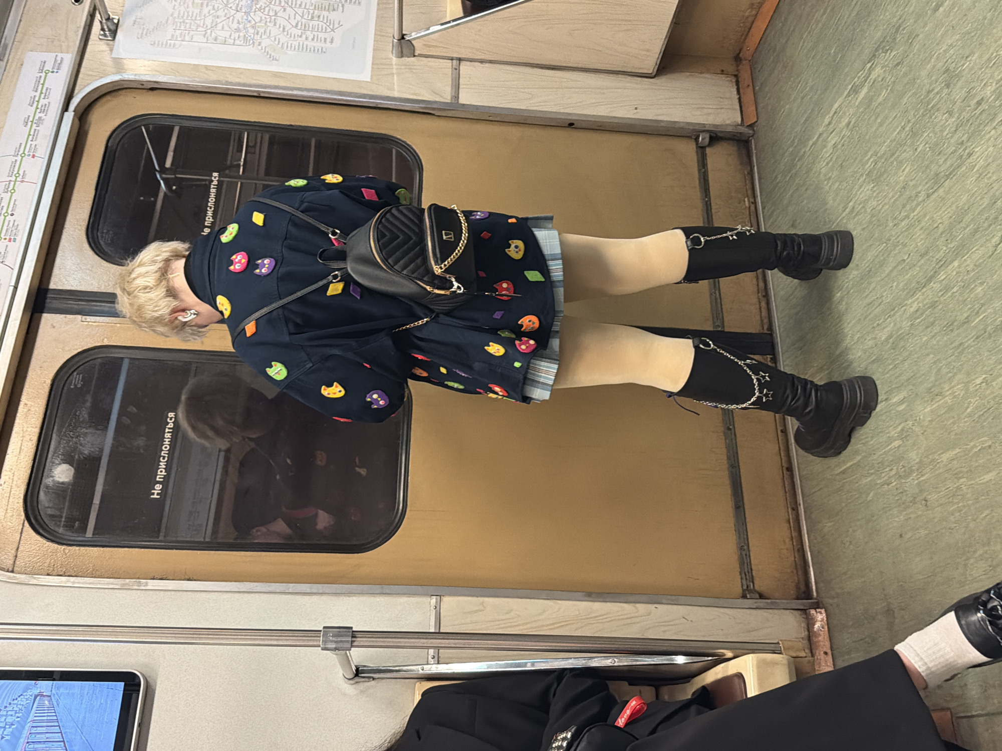 Inside a Moscow Metro train car, a blonde woman in a dark jacket decorated with colorful cat shapes sits sideways in a seat with her legs extended. She wears tan tights and black boots with a chain detail. A black purse hangs on her shoulder. The train car's window is to her left. A subway map is on the wall above the window. A person sits next to her, wearing black pants and one visible shoe with a white sock. The train car's floor is green.