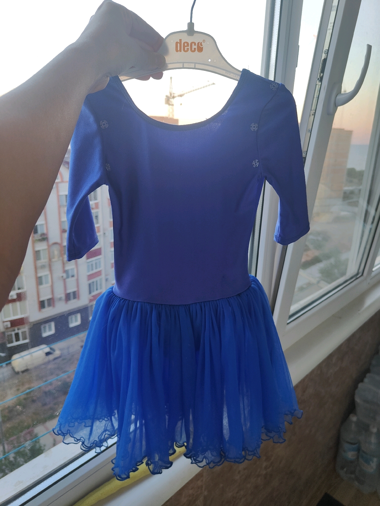 A blue, short-sleeved dress with a gathered tulle skirt is displayed on a white hanger, held by a person's hand, in front of a window. The dress has a round neckline and is decorated with small, silver, star-shaped embellishments on the shoulders. The background through the window shows a residential building, a parked white van, and a construction crane, indicative of an urban setting. The scene appears to be an apartment or room in the city of Chornomorsk, Ukraine, with the window offering a view of the exterior. The lighting suggests it is either early morning or late afternoon.