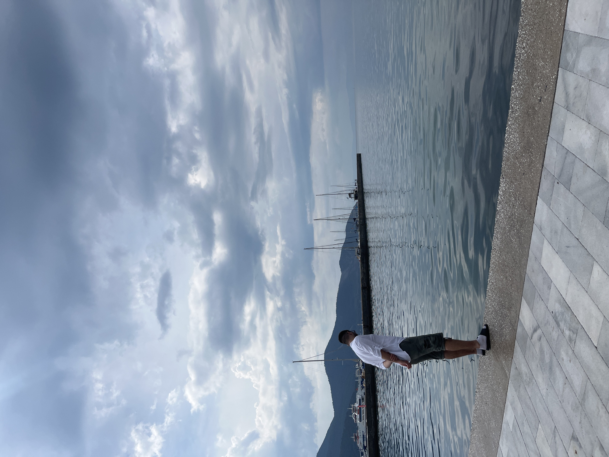 A lone individual stands with their back to the viewer, facing a body of water. The person is wearing a white short-sleeved button-down shirt, camouflage-patterned shorts, white socks, and dark sandals. Their hands are clasped behind their back.
The scene is a waterfront promenade, paved with large grey and white rectangular tiles. Adjacent to the paved area is a stone border, followed by a darker grey walkway, and then the calm, rippling water of a harbor.
In the middle ground, a long pier or breakwater extends into the water, lined with several sailboats with tall masts. Beyond the pier, a range of mountains forms the backdrop, receding into the distance. A ferryboat is docked on the left side of the pier.
The sky above is filled with a mix of white and grey clouds, with some shafts of sunlight breaking through, suggesting an overcast or partially cloudy day. The overall atmosphere is serene and contemplative. The location appears to be a harbor in the city of Thassos, Greece, given the architectural style and the presence of sailboats and a ferry.