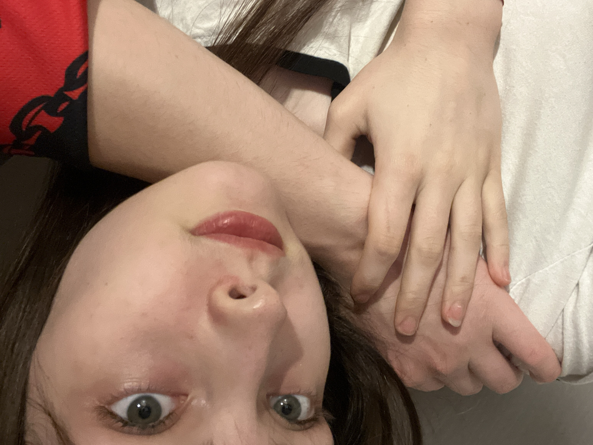 A close-up selfie captures a person, likely female, lying down and looking directly at the camera. The subject has fair skin, long dark hair, and wide, light-colored eyes, possibly green or hazel, with a direct gaze. Her lips are lightly glossed with a reddish tint and slightly parted. Fine hair is visible on her arms and face, and a few small blemishes or freckles are present, indicating a natural appearance.
The person's head is tilted to the side, occupying the lower left portion of the frame. On the left side of the image, a red fabric with a black pattern (possibly part of a garment) is visible. On the right, a white or cream-colored soft fabric, resembling bedding or a plain shirt, is present. Two hands are interlocked or one is holding the other's arm, visible on the right side of the frame, blending with the white fabric. The hands also show natural, unpolished nails and some fine hair.
The lighting is soft and diffused, suggesting an indoor setting. The composition creates an intimate, personal perspective, characteristic of a selfie. No specific location cues for Saint Petersburg, Russia, are discernible from the image itself. There is no visible text.