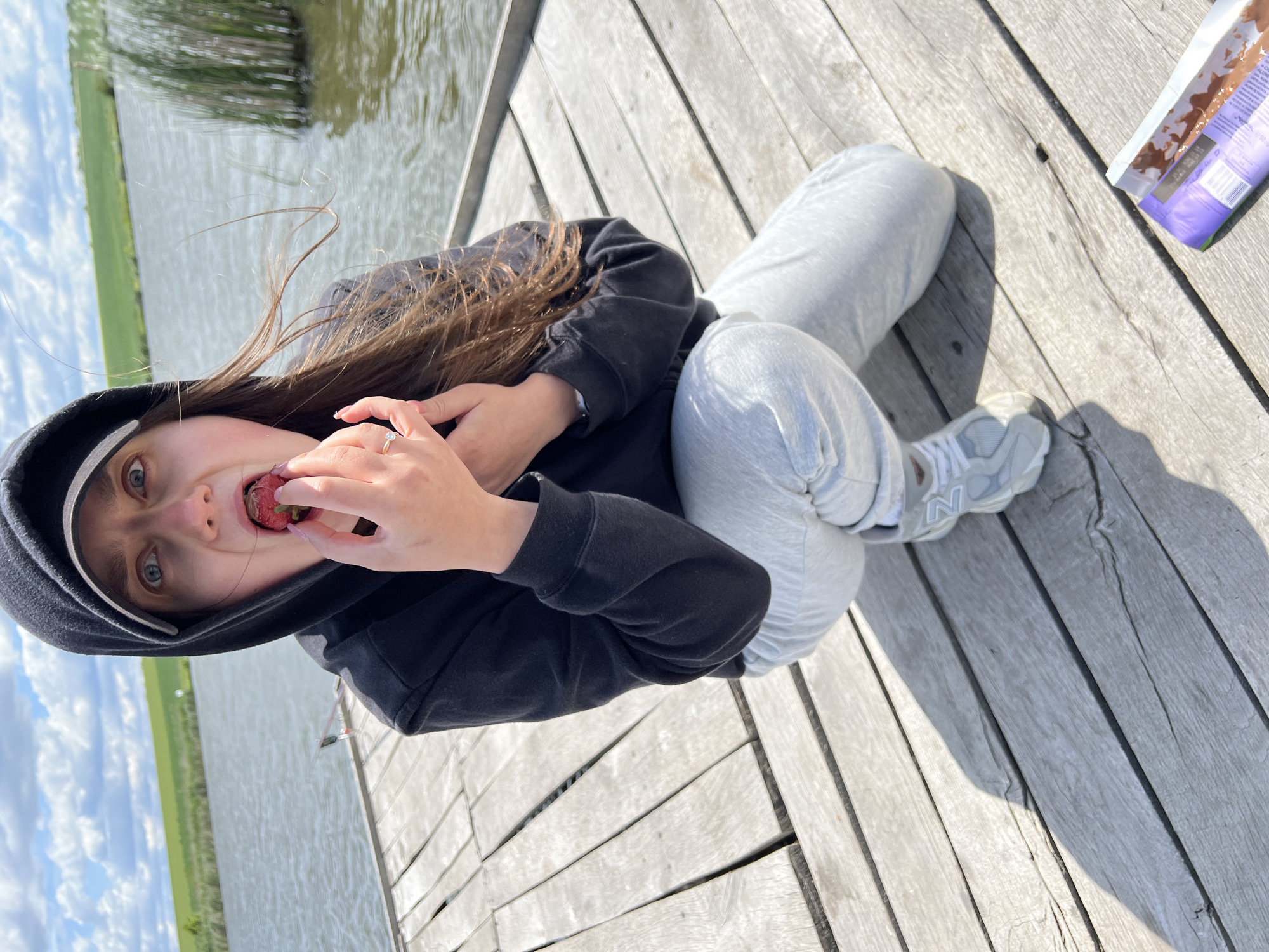 A young woman sits cross-legged on a wooden dock, likely constructed for recreational use. The dock appears to be built on a lake, as water is visible in the background. She is wearing a black hooded sweatshirt, gray sweatpants, and gray sneakers. She is holding a strawberry in her hand and is in the process of eating it. Next to her is a box of chocolate bars. The geographical location is not determinable based on the image.