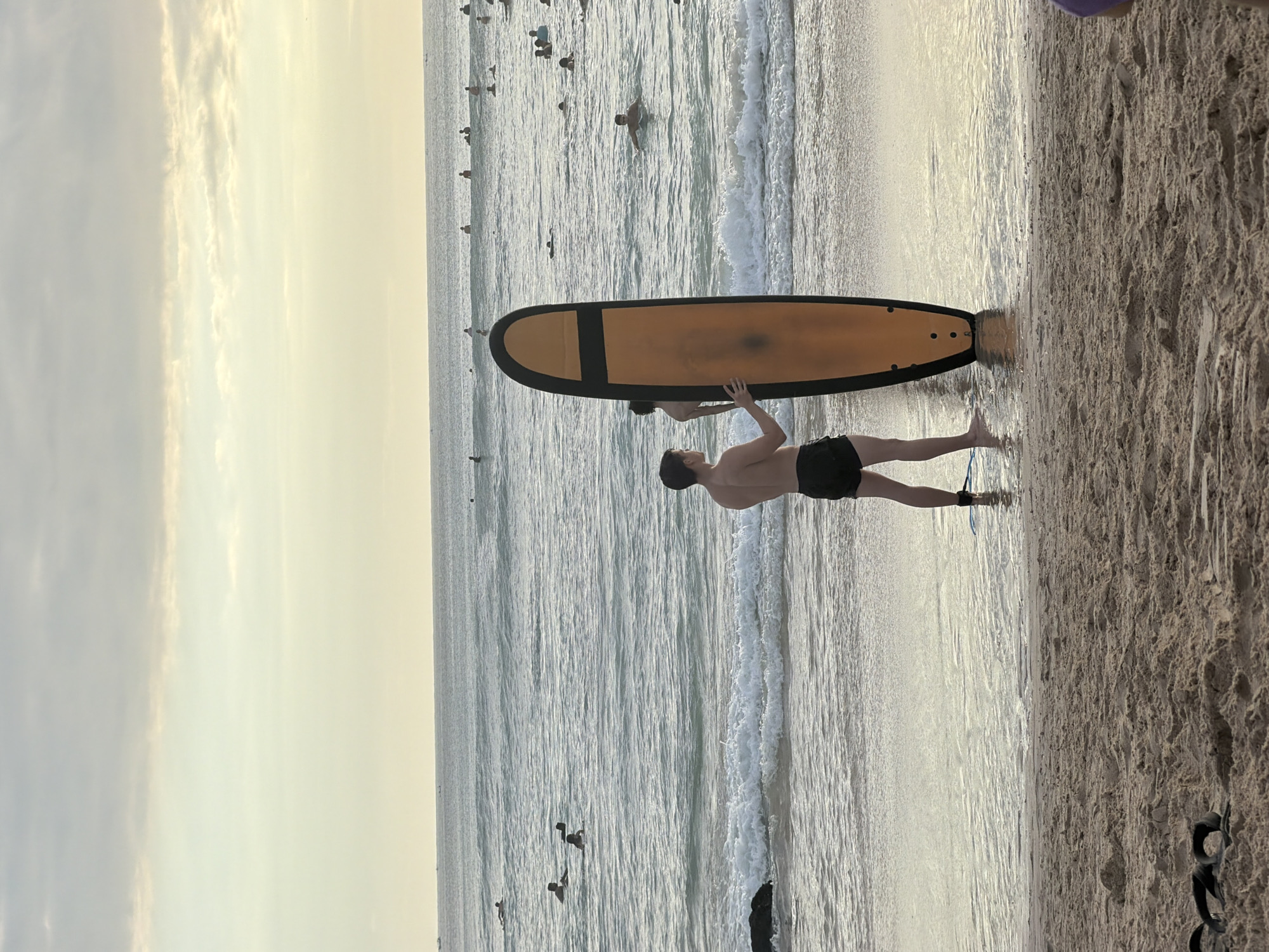 A shirtless man wearing dark swimming trunks stands on a sandy beach near the water's edge, holding a surfboard. The surfboard is mainly orange with a black outline. A second person is bending over the surfboard, presumably assisting the man. In the background, the ocean is visible with several people swimming or surfing. The sky is overcast. The location is likely near Kuta Selatan, Indonesia, due to the beach environment.
