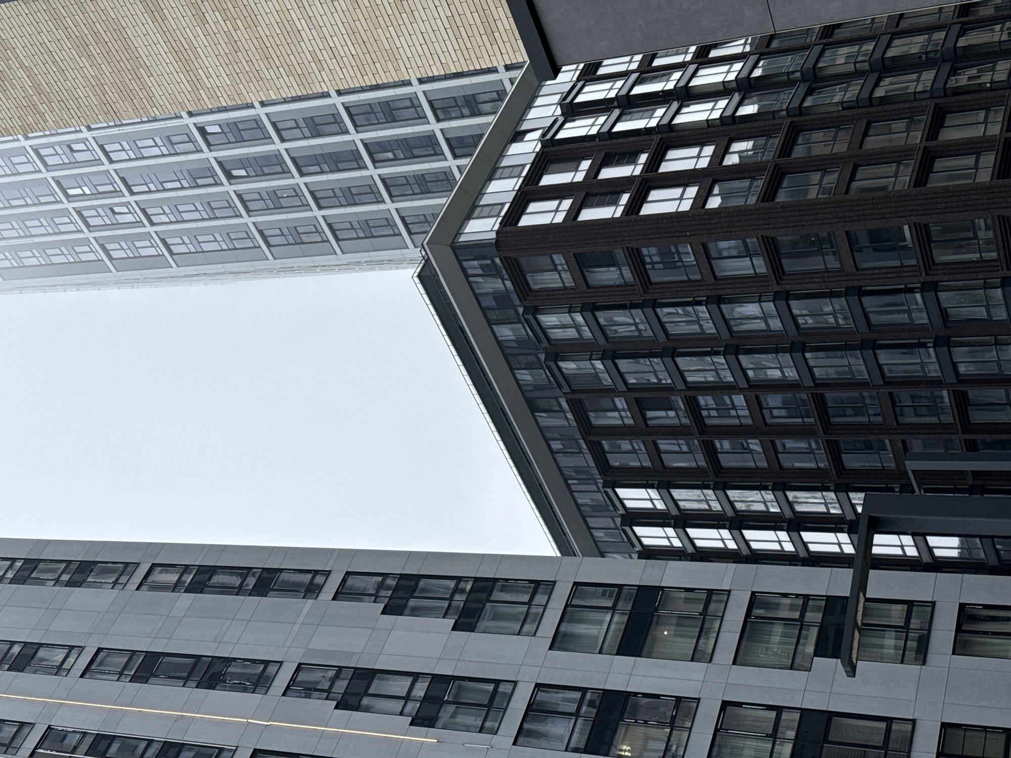 The image shows a low-angle view of several modern buildings, with a patch of sky visible between them. The buildings feature grid-like patterns of windows, some reflecting the sky. The building on the right has a darker facade, while the one on the bottom is gray. The building on the top left has a brick-like appearance on top and gray windows below.
The image does not contain information on whether it is located in Moscow, Russia. The image does not contain information on people, subjects, objects, actions, and interactions.