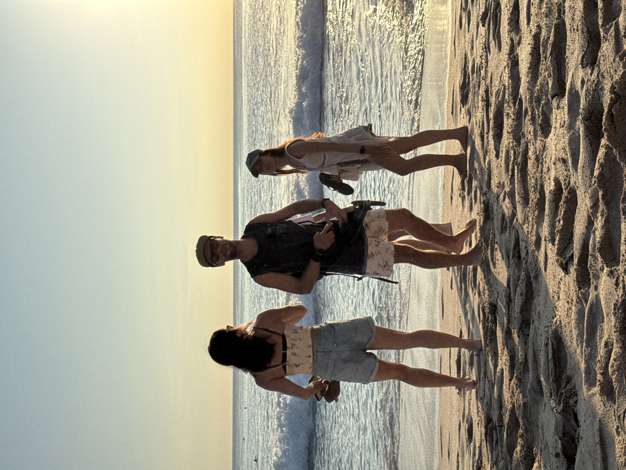 At a beach in Kuta Selatan, Indonesia, three individuals are walking on the sand near the water's edge. One individual wears a dark tank top and floral shorts. The second individual wears a light dress and blue hat. The third individual wears a floral top and blue shorts. The beach is sandy and the water is calm.