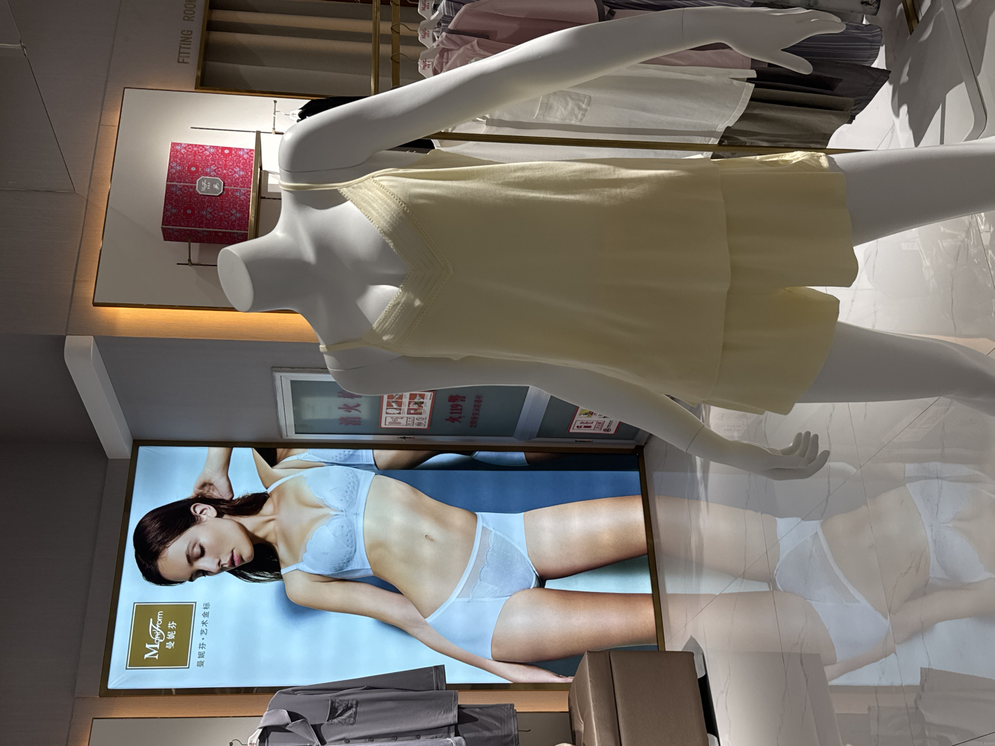 The image shows a retail display inside a store, likely in Linghe, China, as indicated by the signage. In the foreground, a white mannequin is dressed in a light yellow lace-trimmed camisole and matching shorts. Behind the mannequin, a large illuminated advertisement displays a woman wearing a light blue bra and matching underwear. To the right of the advertisement, a sign reads "FITTING ROOM" in English and what appears to be Chinese characters. Further to the right, a rack of clothing is visible, and a closed fitting room door can be seen. The store environment features polished marble floors, and a pink gift box is displayed on a shelf in the background. The overall scene depicts a lingerie or women's apparel store display.