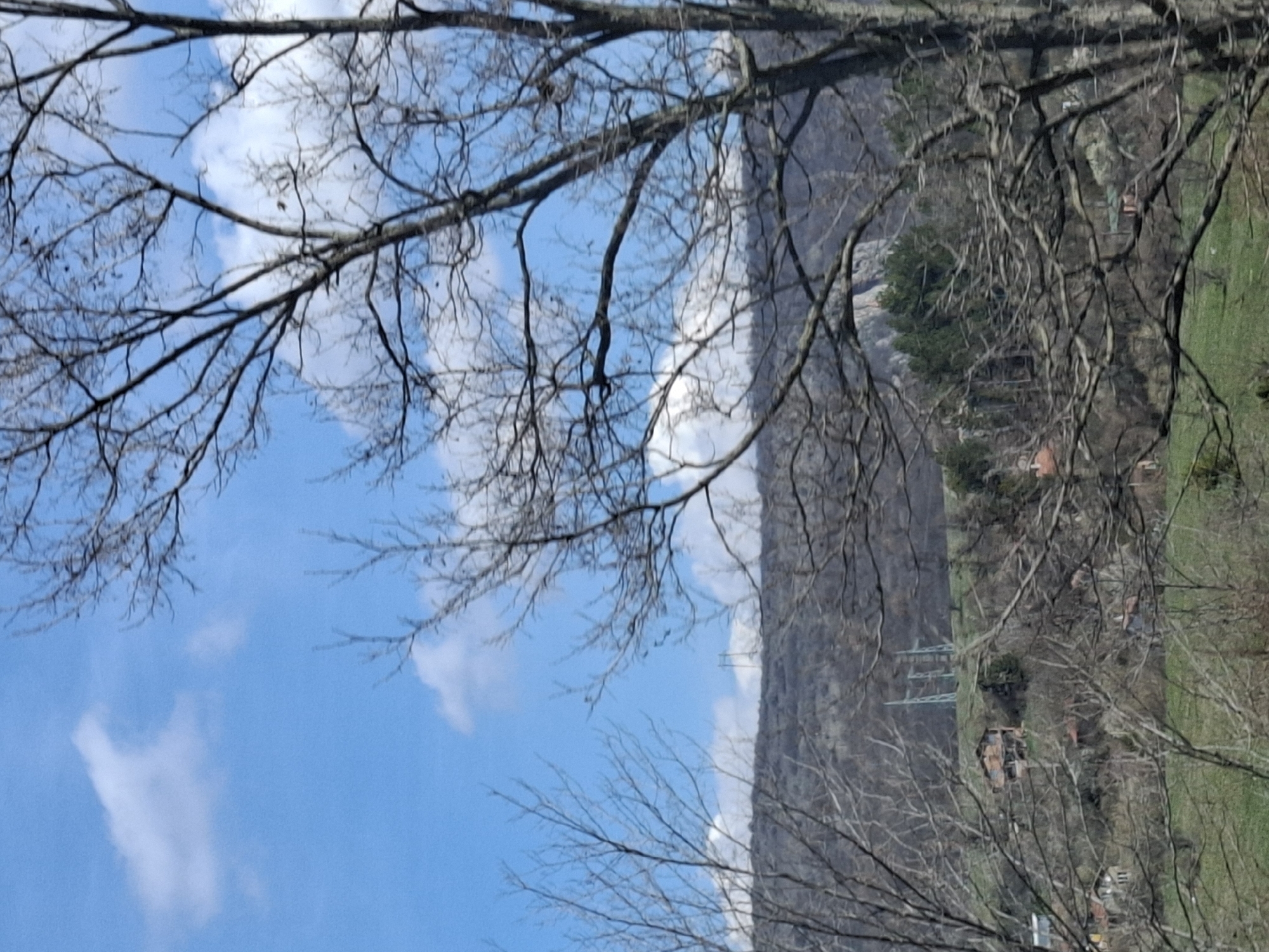 The image depicts a scene in Gradets, Bulgaria, featuring a mountain range with a visible cliff face running along the middle of the frame. The lower slopes of the mountain are partially forested, with occasional buildings and power lines visible. In the foreground, bare tree branches frame the mountain and a blue sky with scattered white clouds. The perspective is slightly angled, capturing both the sky and the terrain below.