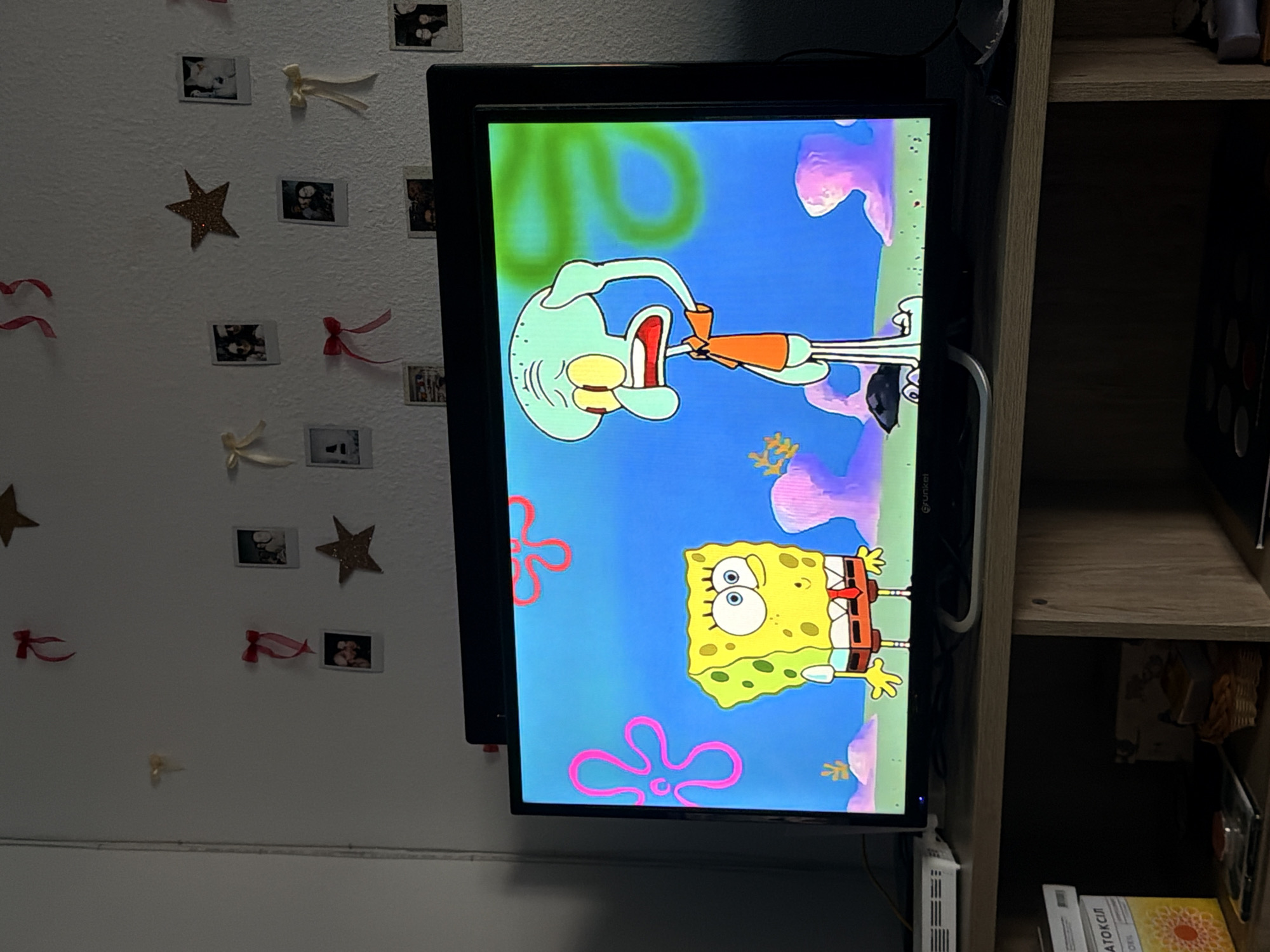 A television set displays a scene from the animated series SpongeBob SquarePants, featuring SpongeBob and Squidward. The TV is positioned on a wooden media console. The background of the scene shows a vibrant blue underwater environment with pink sea anemones. The wall above the media console is decorated with various small framed photographs, glittery stars, and decorative bows. The overall setting appears to be a domestic interior, possibly a living room or bedroom, in Castillo Bajo-conejito, Spain.