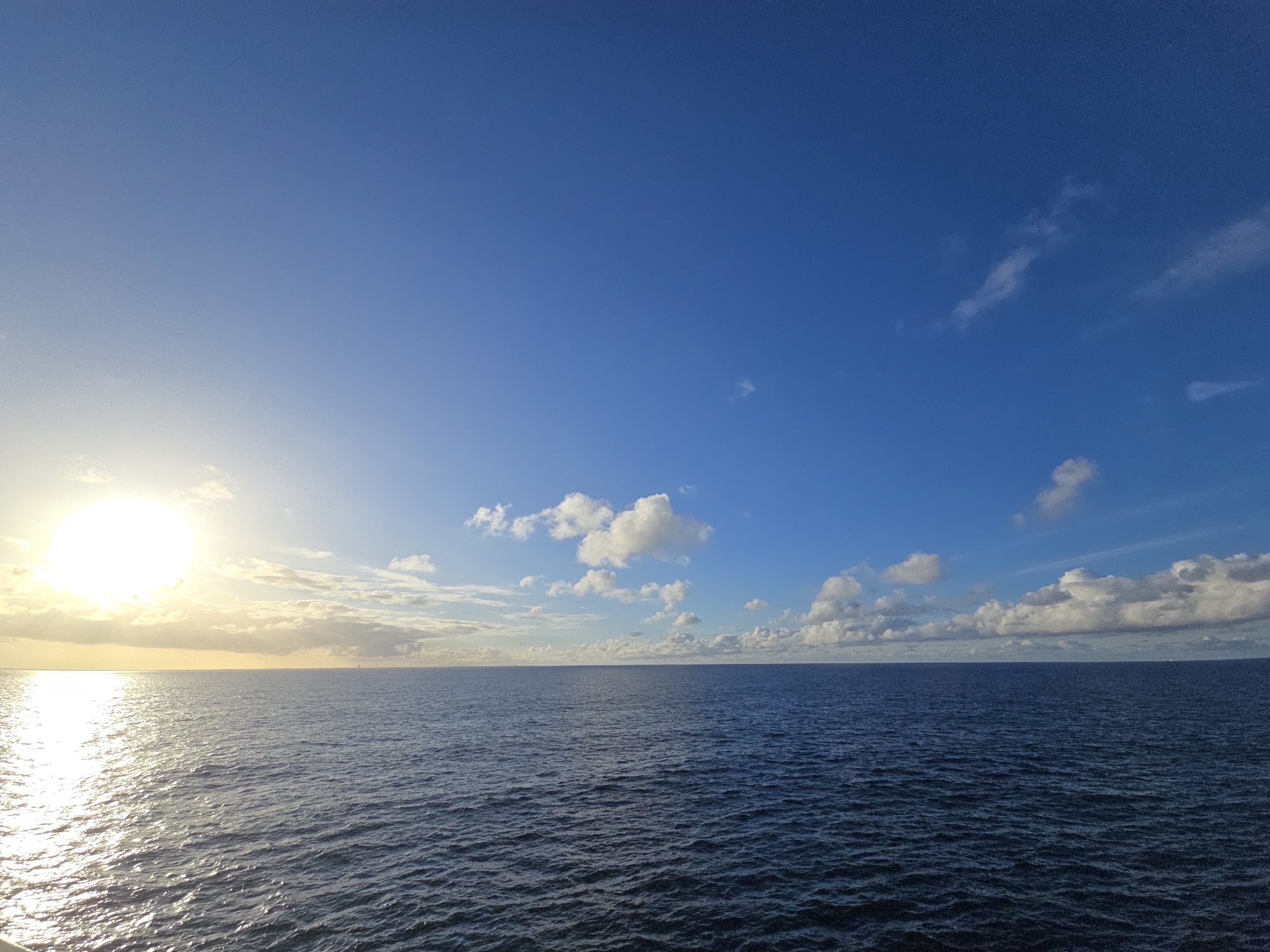 The image shows a view of the ocean under a blue sky with scattered clouds. The sun is positioned low on the horizon to the left, reflecting brightly on the water's surface. The ocean appears dark blue and choppy, with small waves visible. The horizon line is relatively straight and even. There are no people or objects visible in the image other than the sky, water, and sunlight. The location is unknown.