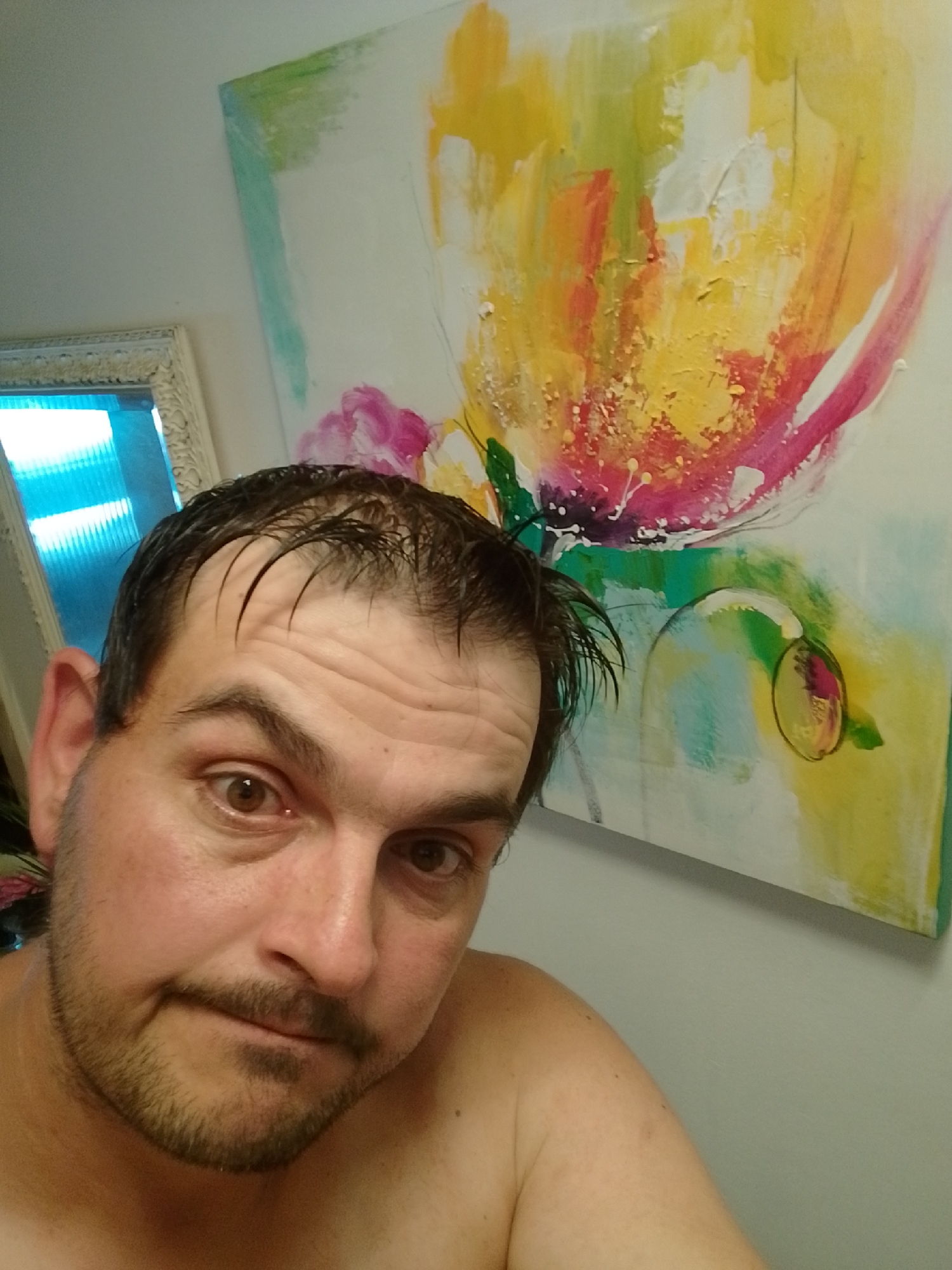 The image is a close-up, front-facing selfie of a shirtless man with short, dark, wet hair and a light beard. He has brown eyes and a neutral expression. He is positioned in the foreground, with his face occupying a significant portion of the frame. Behind him, to the left, is a decorative mirror with an ornate frame, reflecting blue light. To the right of the mirror and behind the man's head is a large, abstract painting featuring bold strokes of yellow, pink, red, and green. The background wall is a light gray. The scene appears to be indoors, likely in a residential setting such as a bathroom or bedroom. No specific location in Wedel, Germany, can be identified from the image itself.
