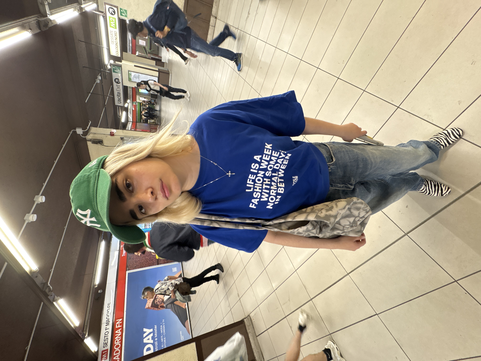 A young person with blonde hair and a green baseball cap is featured prominently in the foreground, looking directly at the camera. They are wearing a bright blue t-shirt with white text that reads "LIFE IS A FASHION WEEK WITH SOME NORMAL DAY IN BETWEEN," light blue jeans, and distinctive black and white zebra-print socks. A silver cross necklace is visible, and they carry a patterned tote bag over their shoulder.
The scene is set in an underground subway station, likely in Milan, Italy, as indicated by various signs. The floor is covered with light-colored square tiles and dark grout lines. Artificial fluorescent lighting strips illuminate the space from the ceiling.
In the background, several other individuals are blurred due to motion, walking through the station. Visible signs include "M1 SESTO 1º MAGGIO ES" and "CADORNA FN," identifying stops on Milan's Metro Line 1. Other signs direct towards "GESSATE" and "COLOGNO NORD 1" on Metro Line 2, along with a bilingual "Uscita Exit" sign. An advertisement board in the background features text such as "DAY," "strali e Master," and "AI ARTE MODA DITORIA CINEMA E TV." The overall activity suggests commuters moving through a busy transit hub. The time of day is not discernible due to the indoor, artificially lit environment.