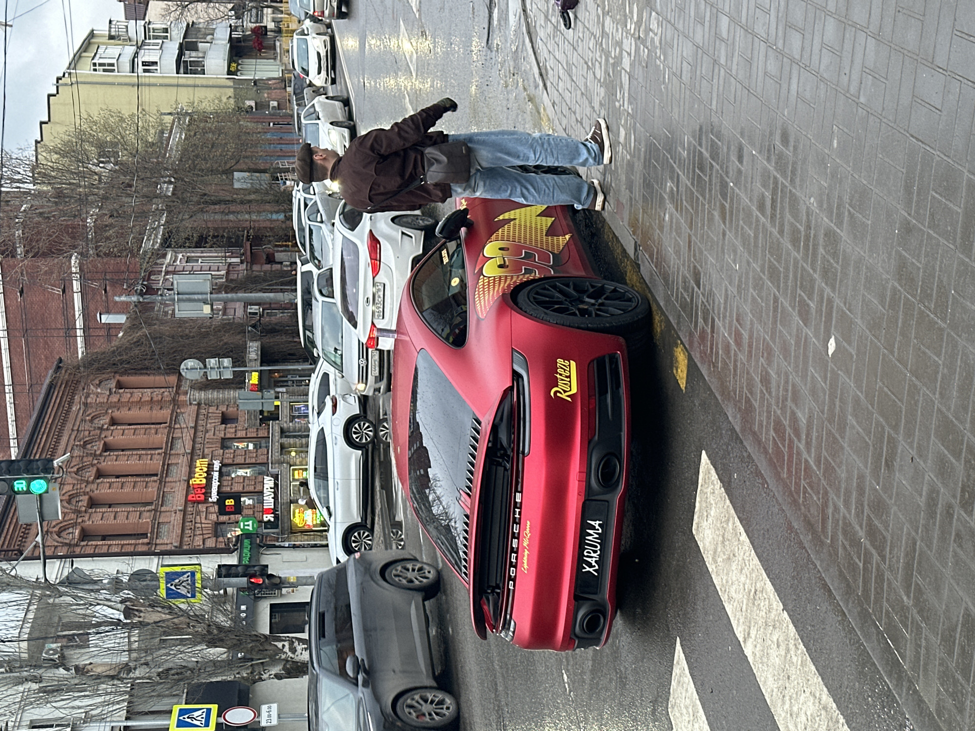 A daytime shot in Rostov-on-Don, Russia, shows a red Porsche car parked on the side of the street with a man standing nearby. The car has a customized design, resembling Lightning McQueen from the movie "Cars". The license plate reads "XARUMA." The man is wearing a brown jacket and jeans, and he appears to be interacting with the car, possibly leaning on it. The street is lined with cars and buildings, and there is pedestrian infrastructure visible in the foreground. The sky is overcast, suggesting a cloudy day. A traffic light pole with a green light is visible on the left. The signs in Russian include "Be Boom" and "I Love Shawarma."