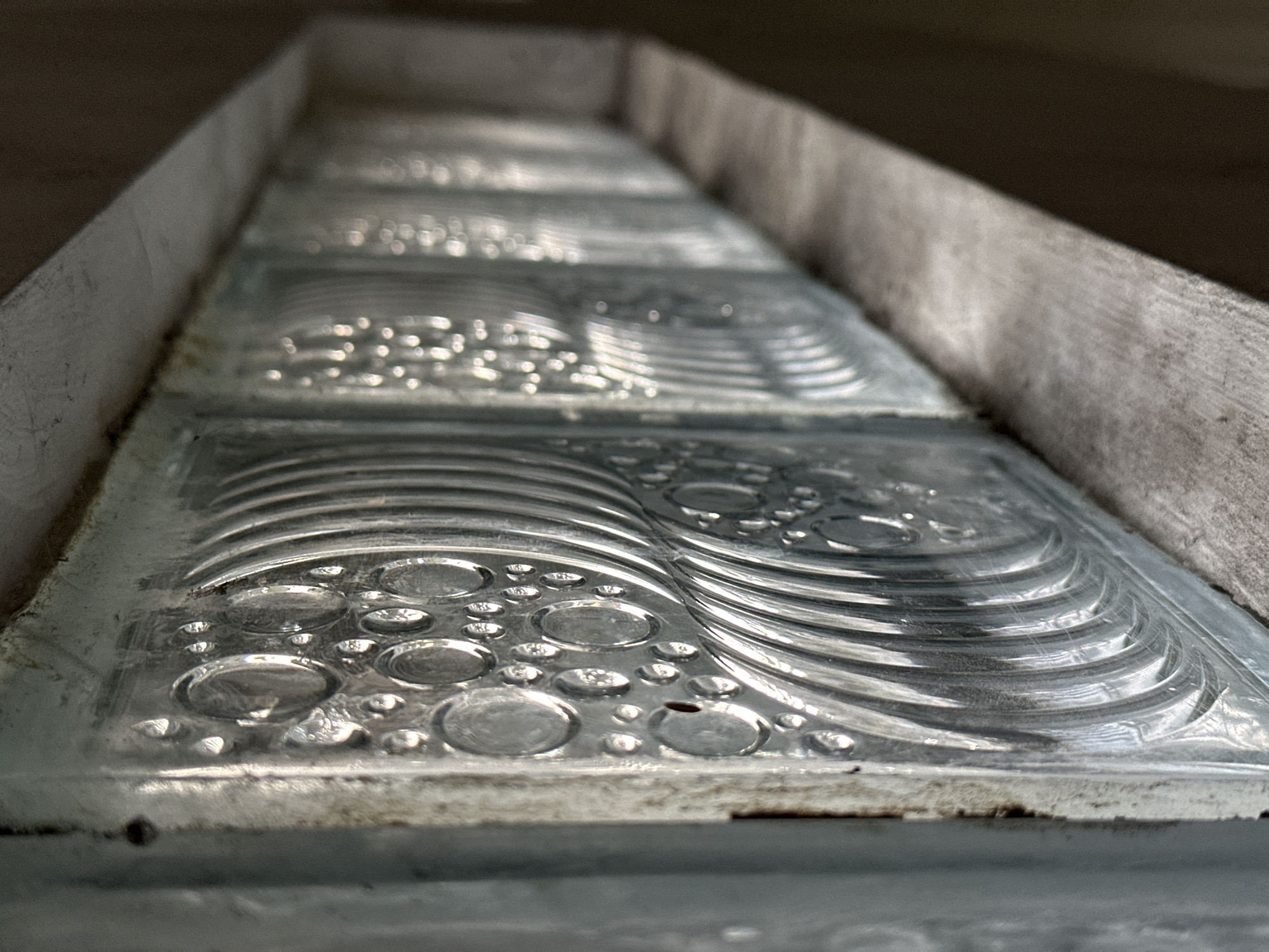 The image is a close-up, eye-level shot of a row of textured glass blocks. The blocks are arranged horizontally and recede into the background, creating a sense of depth. The glass has a pattern of raised circles of various sizes and curved lines. Light reflects off the surfaces of the glass, creating highlights and shadows that emphasize the texture. The blocks appear to be set in a frame or structure made of a greyish, weathered material, possibly concrete or cement, which also lines the ledge below the glass blocks. The overall impression is one of a functional architectural element, possibly a wall or a window, with a focus on the visual qualities of the material itself. There are no people or animals visible in the image. The lighting is natural and appears to be coming from the left side, casting shadows to the right. The environment appears to be outdoors or in a well-lit indoor space with a utilitarian aesthetic. The specific location in Bulakrejo, Indonesia is not discernible from the image itself, as the visual details are solely focused on the architectural elements.