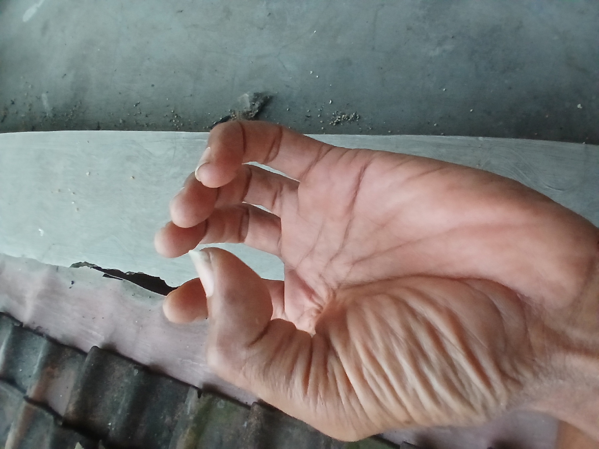 A close-up, high-angle, outdoor shot shows a person's right hand in the foreground, forming the "OK" gesture with their thumb and index finger. The hand is tan with visible wrinkles and veins. The background features a gray, textured concrete surface with scattered small white particles, and on the left, a portion of a dark, textured material resembling roof tiles. This scene appears to be in Tenggarang, Indonesia, possibly near an outdoor area or walkway.